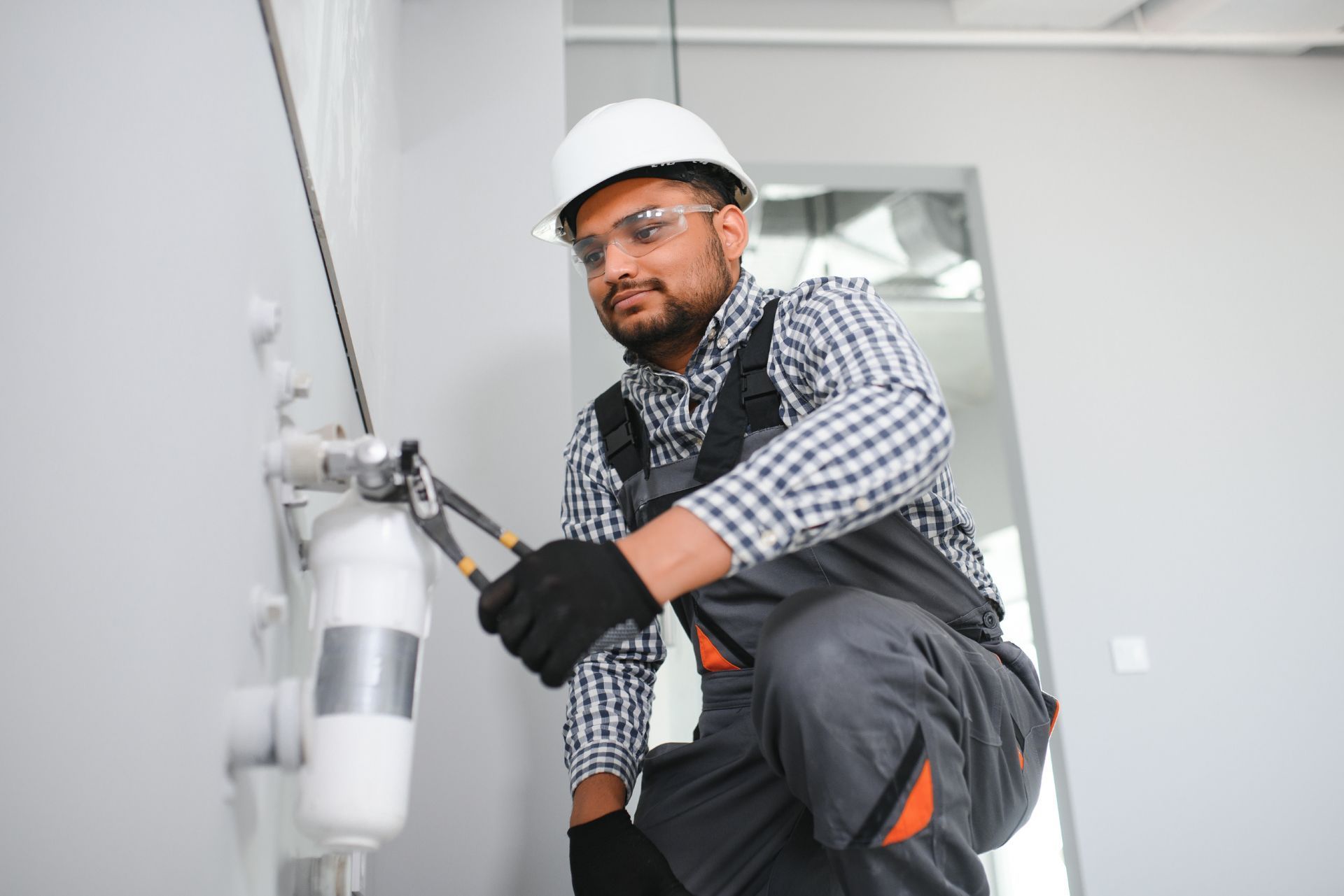 A technician in a white hard hat and safety glasses uses pliers to repair a white cylindrical water filter on a wall.