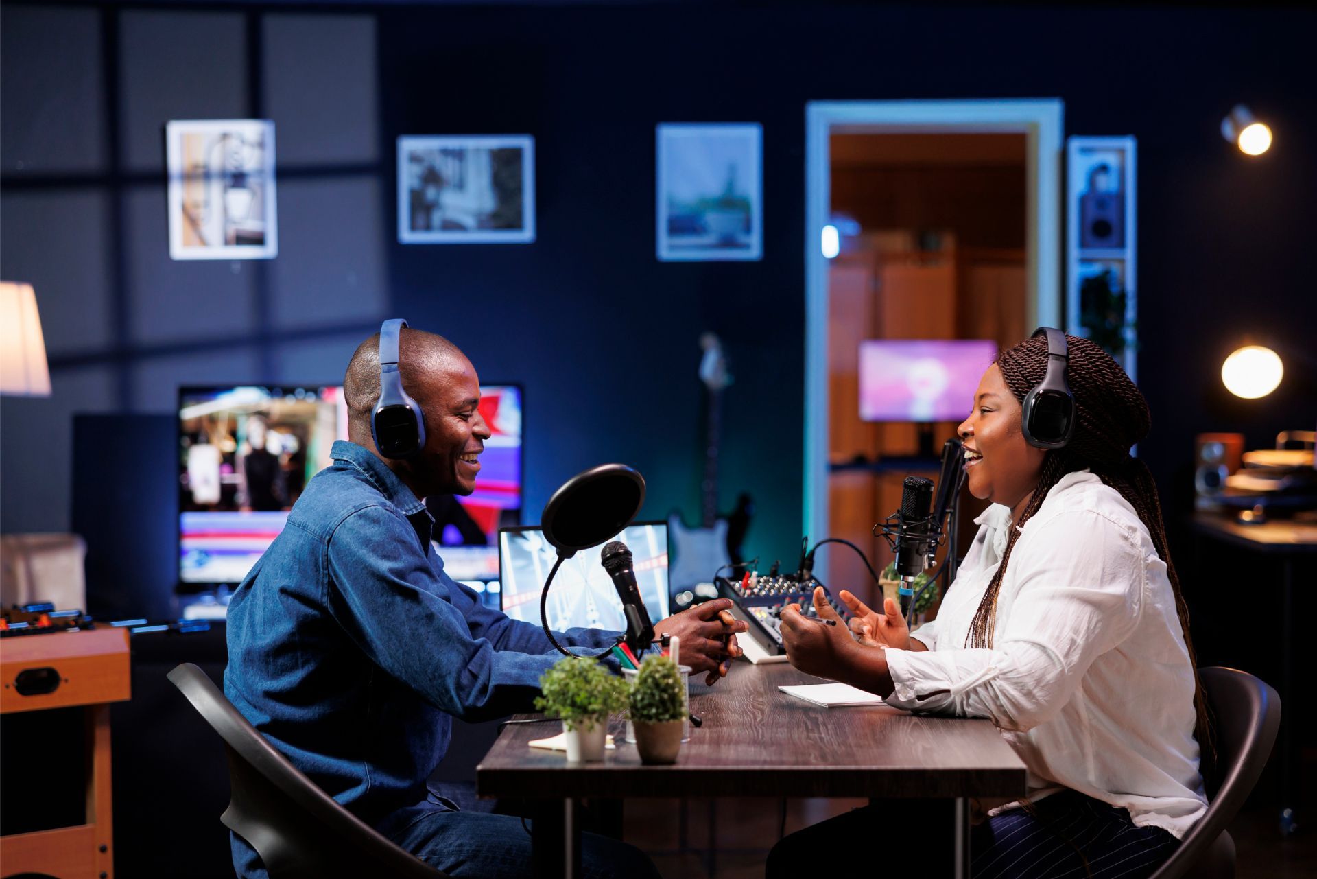 Two people wearing headphones talk across a table into a microphone in a dimly lit home studio.