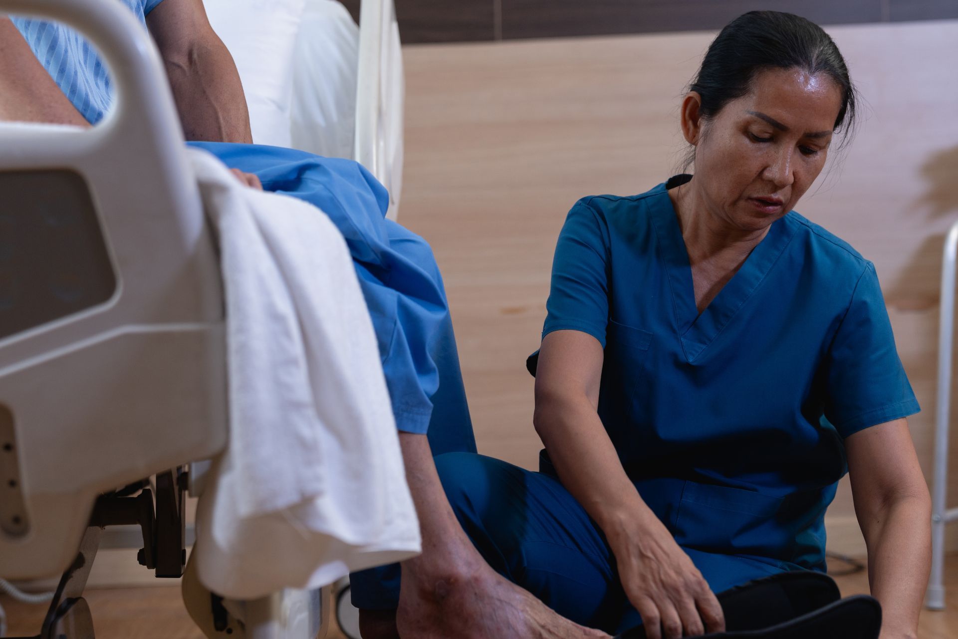 Nurse in blue scrubs sitting beside a hospital bed, looking down while tending to a patient.