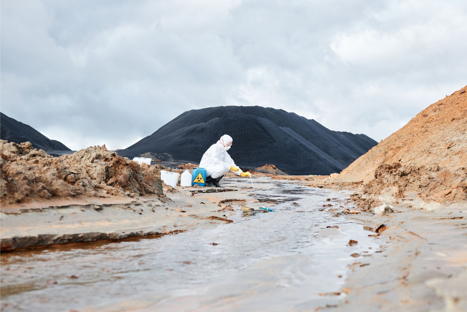Person in white protective suit working in a muddy industrial landscape with hills and cloudy sky