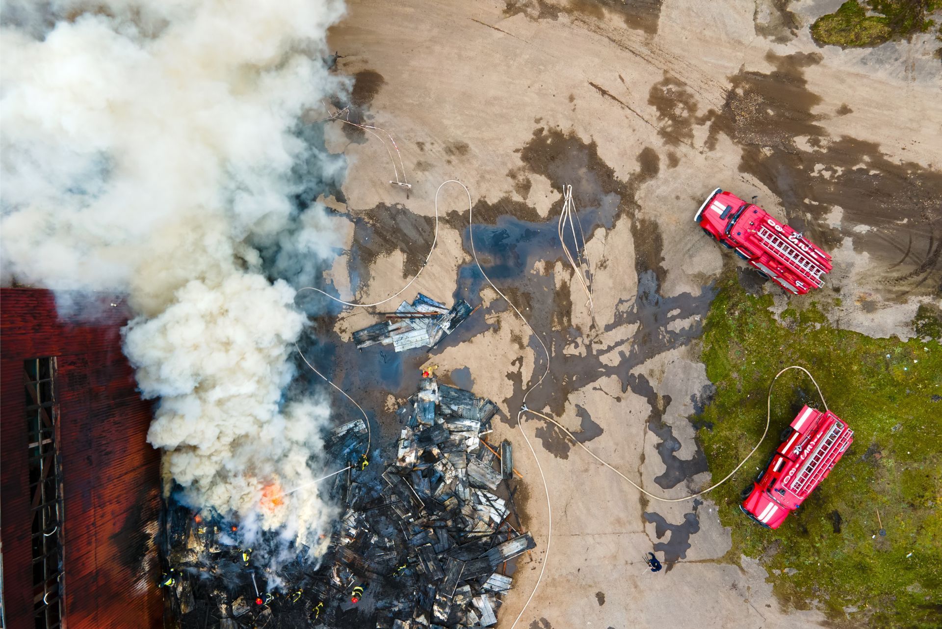 Aerial view of a burning wreck with thick smoke and two red fire trucks on a roadside