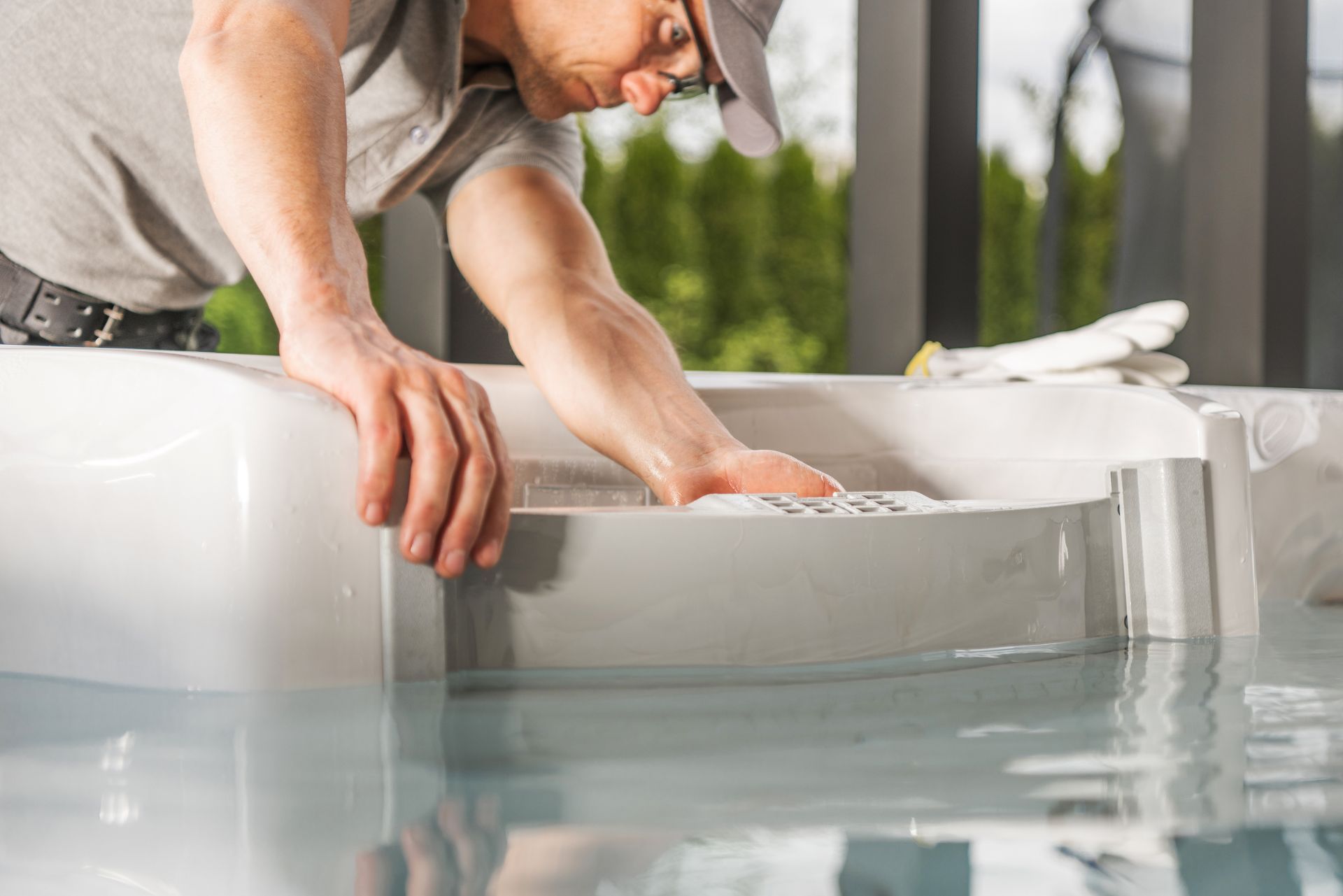A technician in a grey shirt and cap works on a white pool filter system submerged in clear water.
