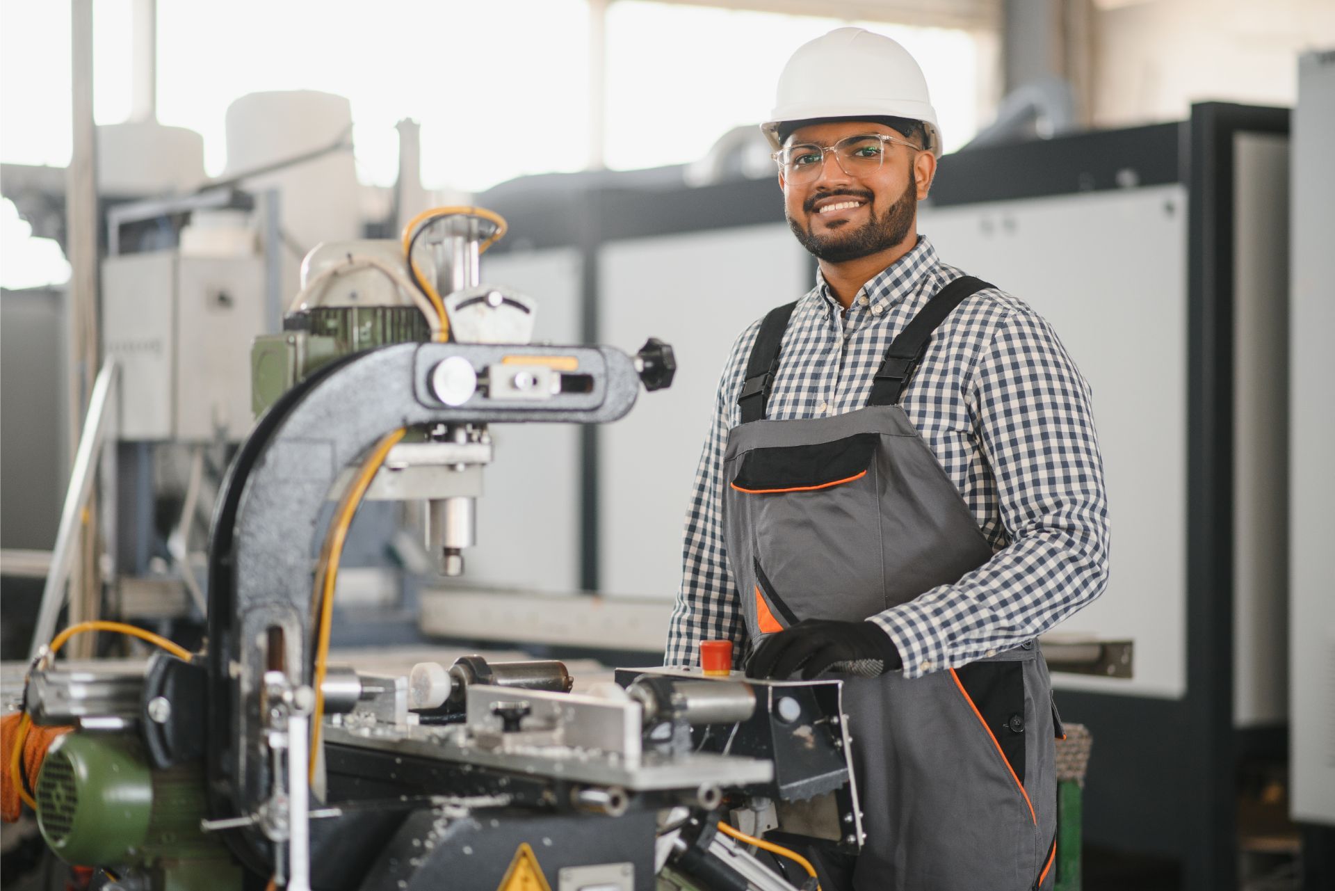 Worker in hard hat and gloves beside industrial machine in a factory, holding a tablet and smiling