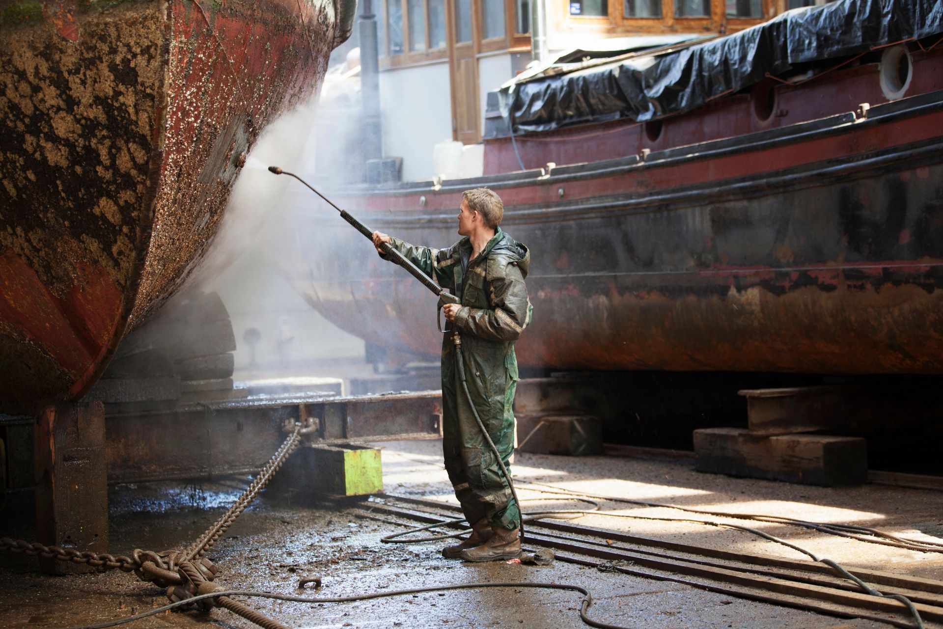 Person pressure-washing a boat in a wet dockyard, with water spraying and another boat in the background