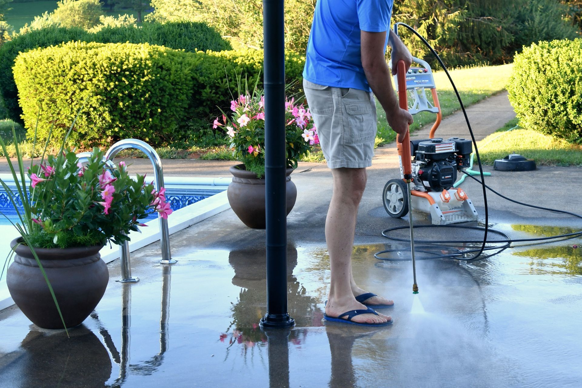 Person washing a driveway with a pressure washer beside potted flowers and a black lamp post