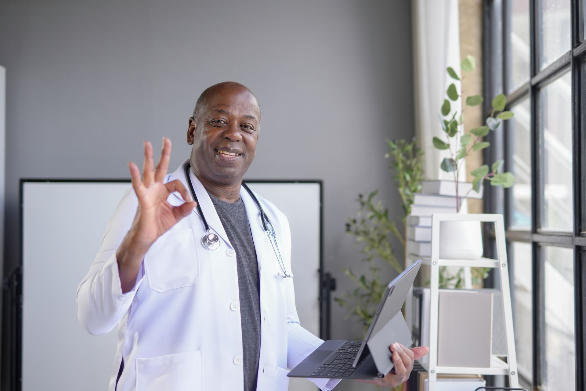Doctor in a white coat holding a clipboard and making an OK sign in a bright office.