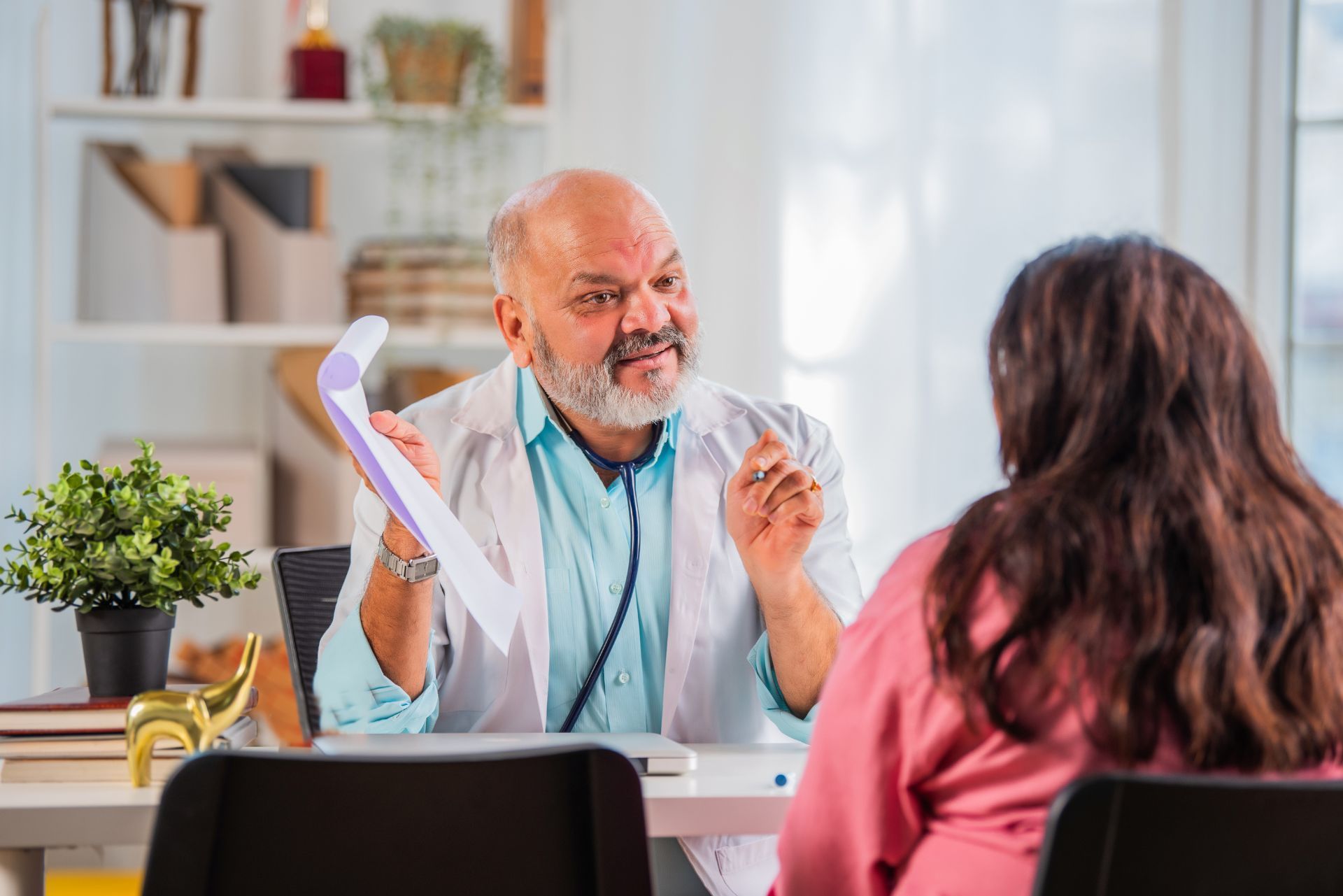 Doctor talking with a patient in a bright office, gesturing while seated behind a desk.
