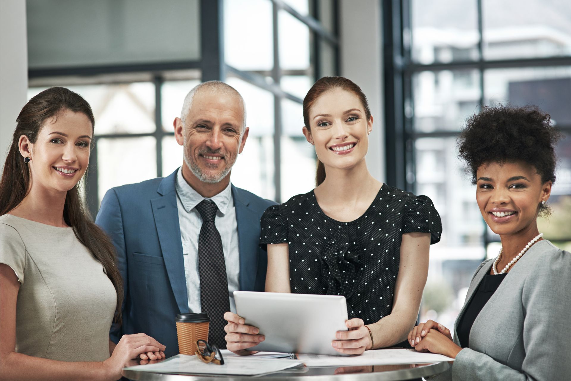 Four coworkers smiling in a modern office, standing behind a table with papers and a laptop.