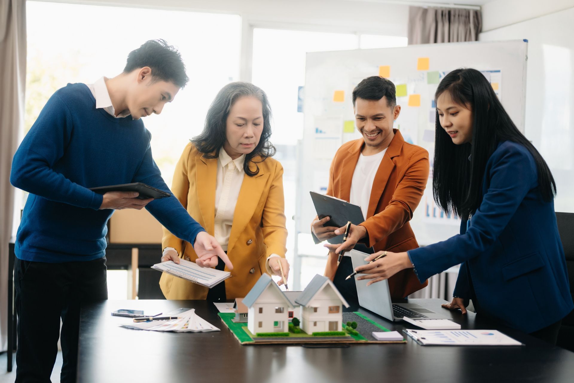 Four people collaborating around a model house at a table, discussing plans in a bright office