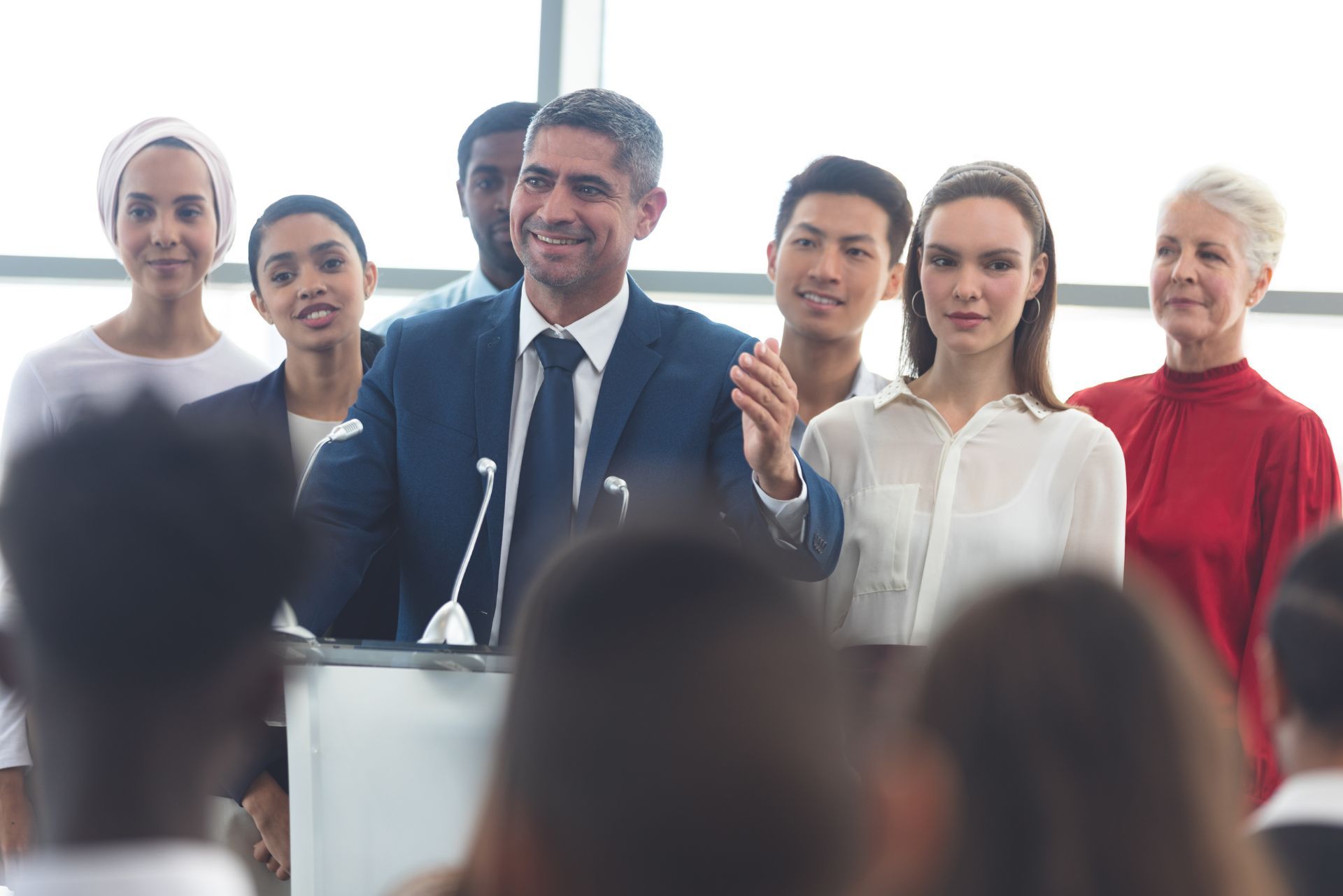 A diverse group stands behind a smiling man in a suit as he speaks at a podium during a presentation.