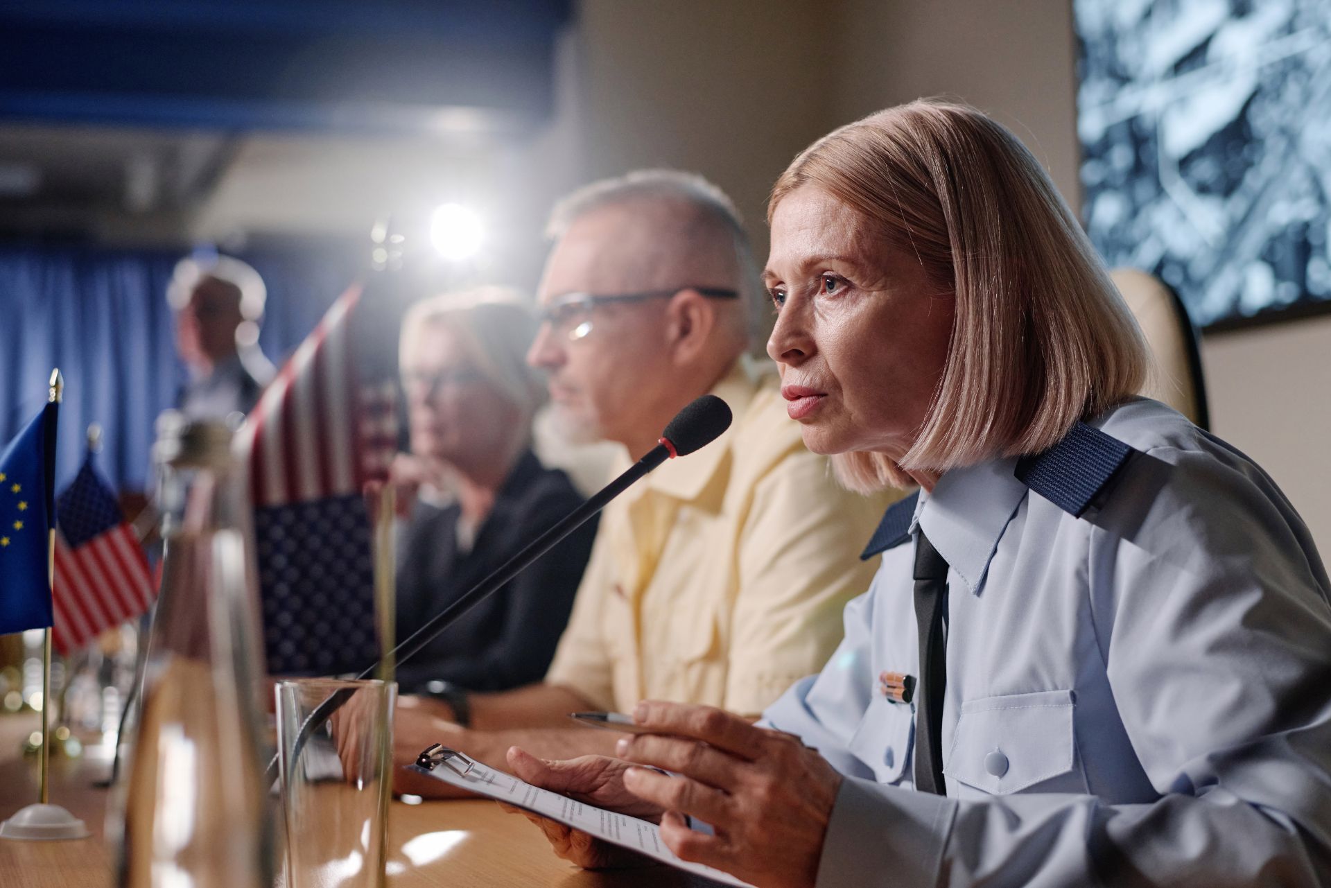 A person in a light blue uniform speaks into a microphone at a table with small flags, next to others at a meeting.