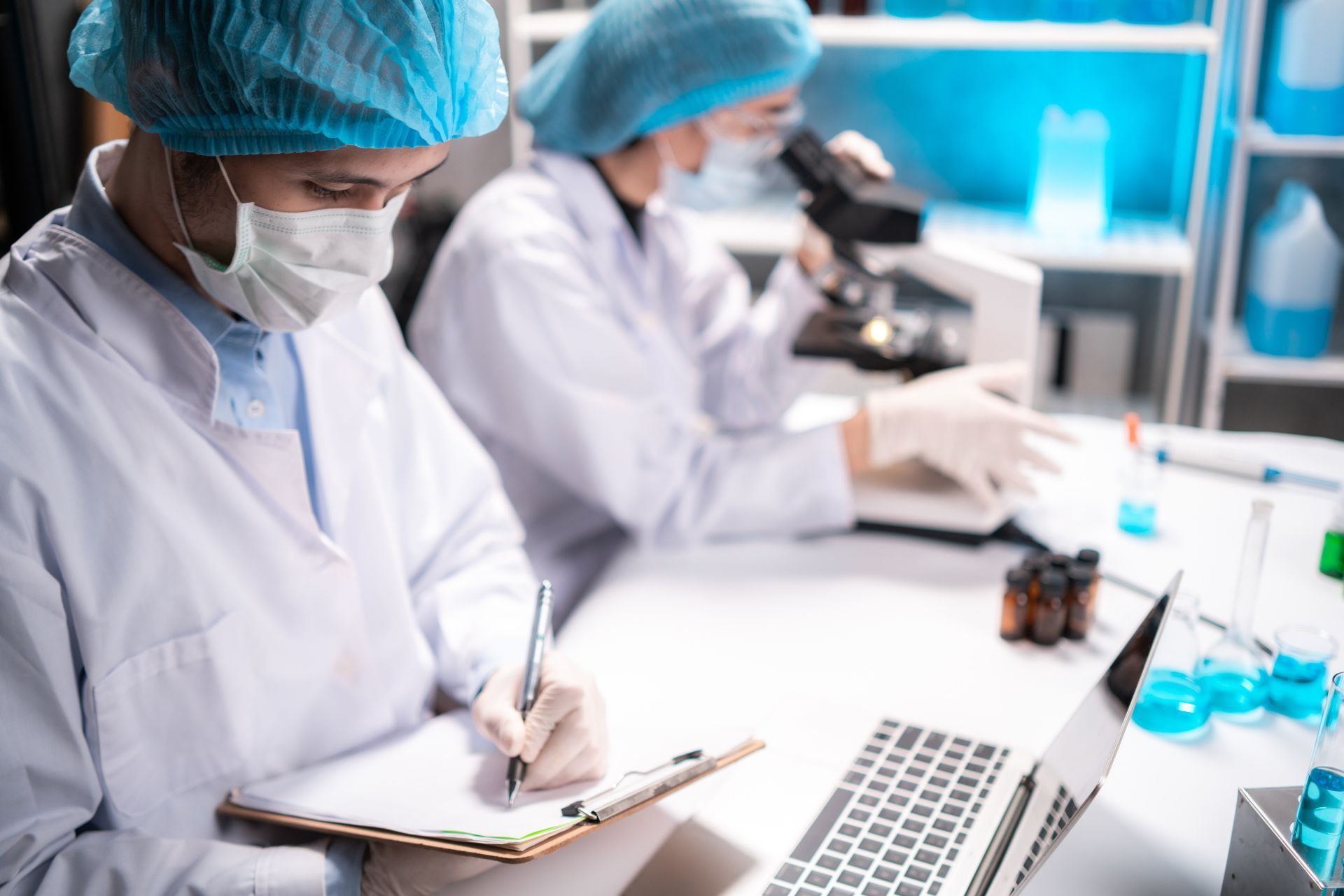 Two scientists in white lab coats and hairnets working in a lab, one taking notes and the other using a microscope.