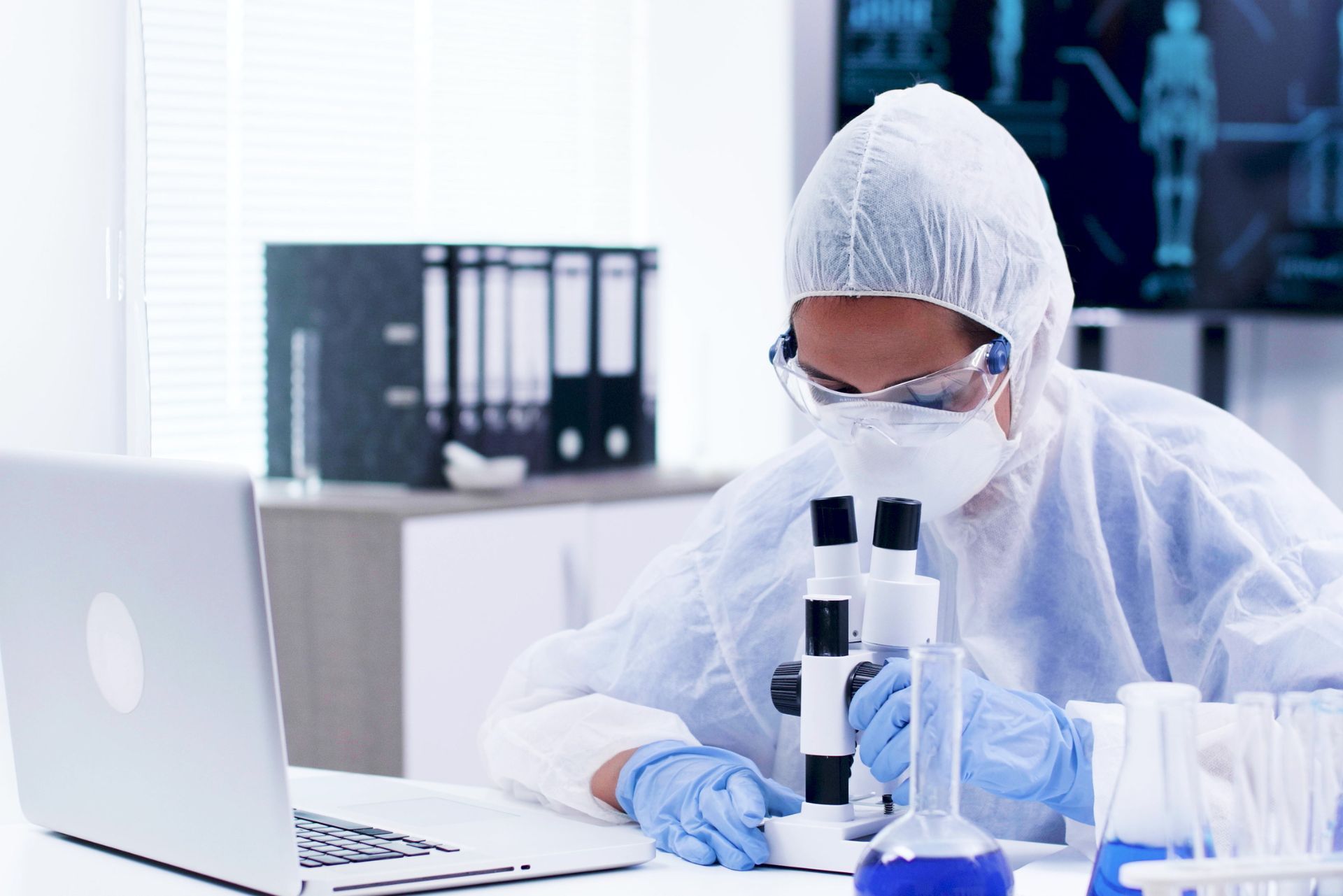A researcher in protective gear, mask, and gloves uses a microscope at a desk with a laptop and lab equipment.