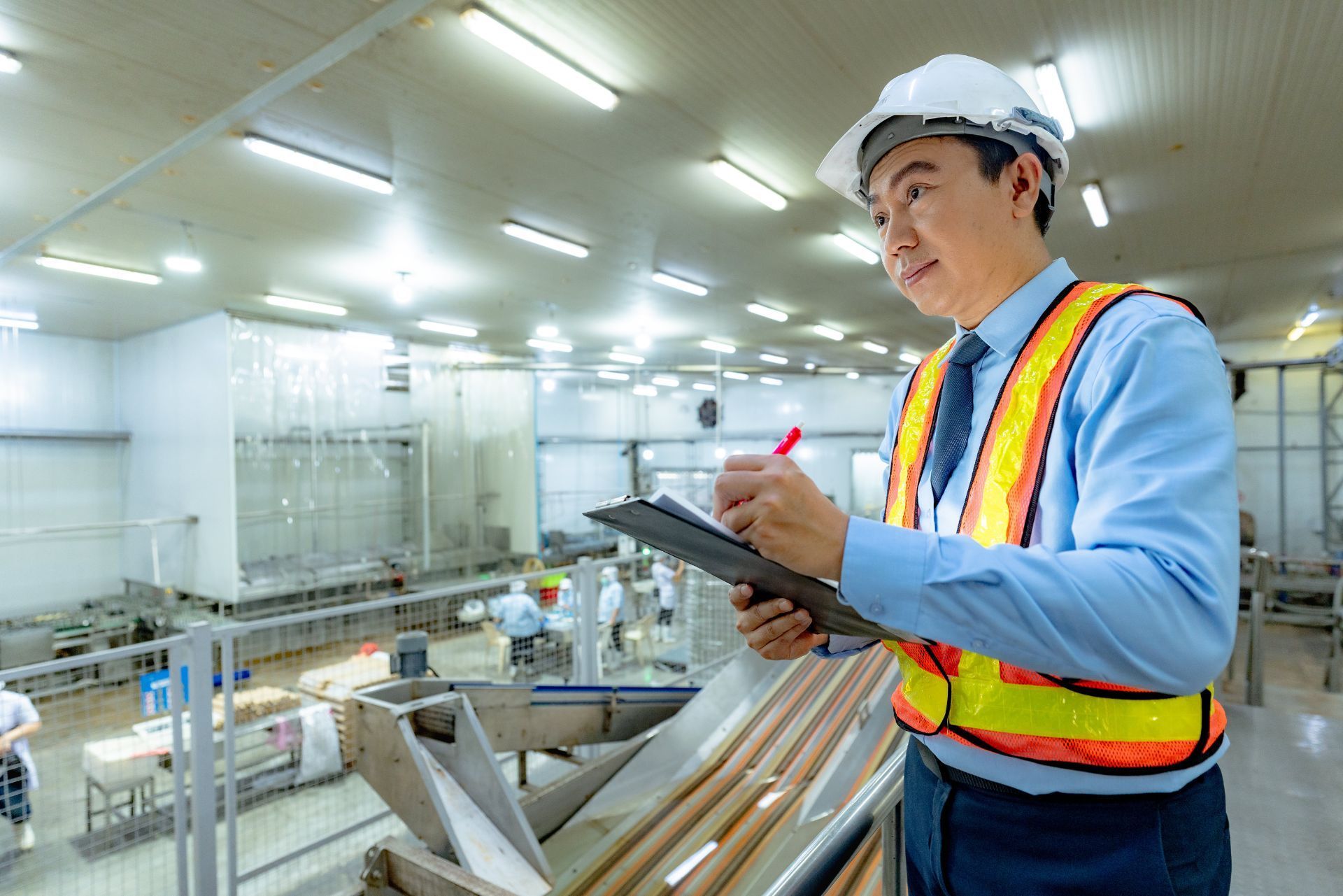 Worker in hard hat and safety vest taking notes in a bright factory aisle