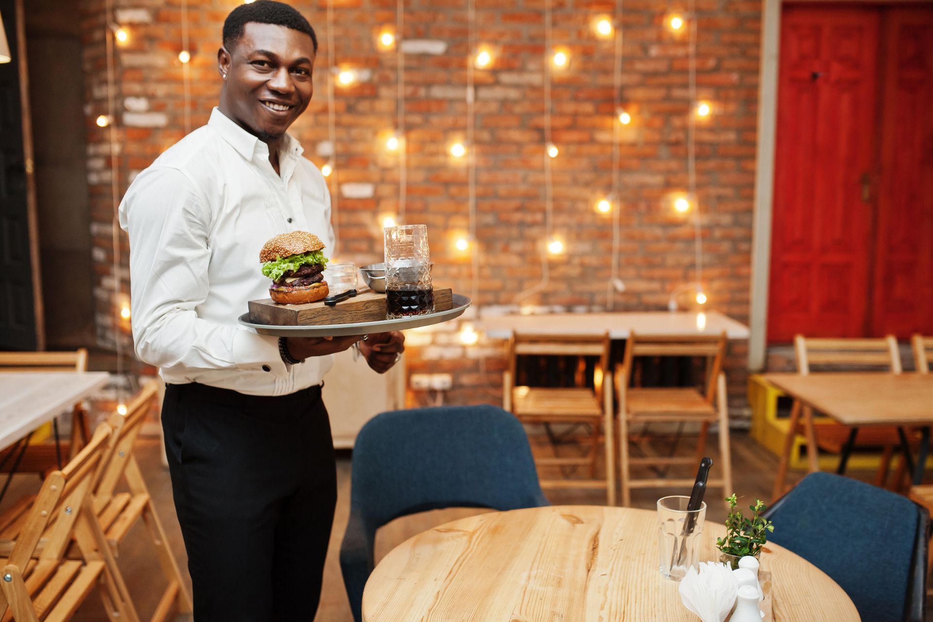 Waiter carrying a tray with burgers and drinks in a warmly lit restaurant.
