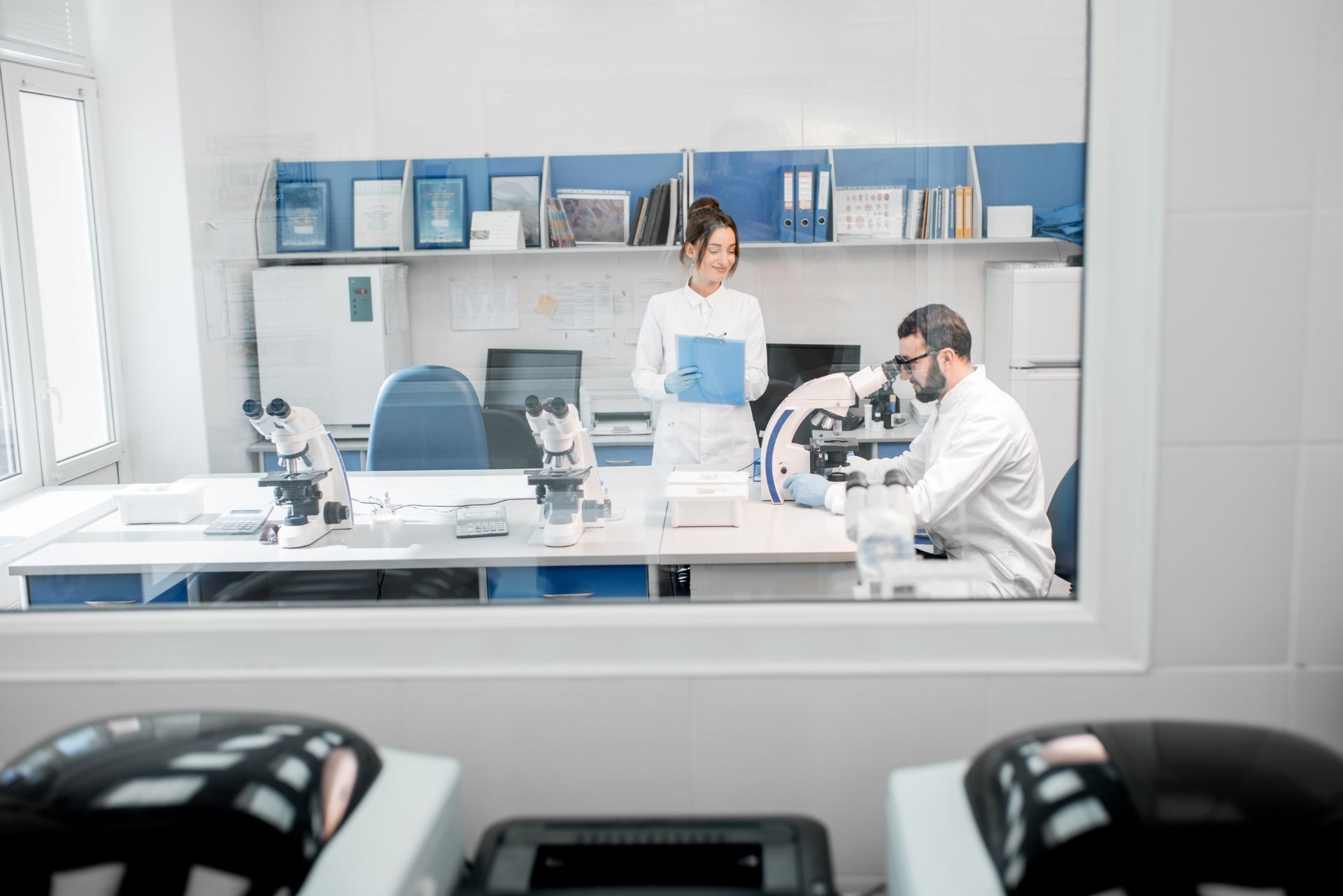 Two researchers in a bright lab, one standing with a tablet and one seated at a microscope.
