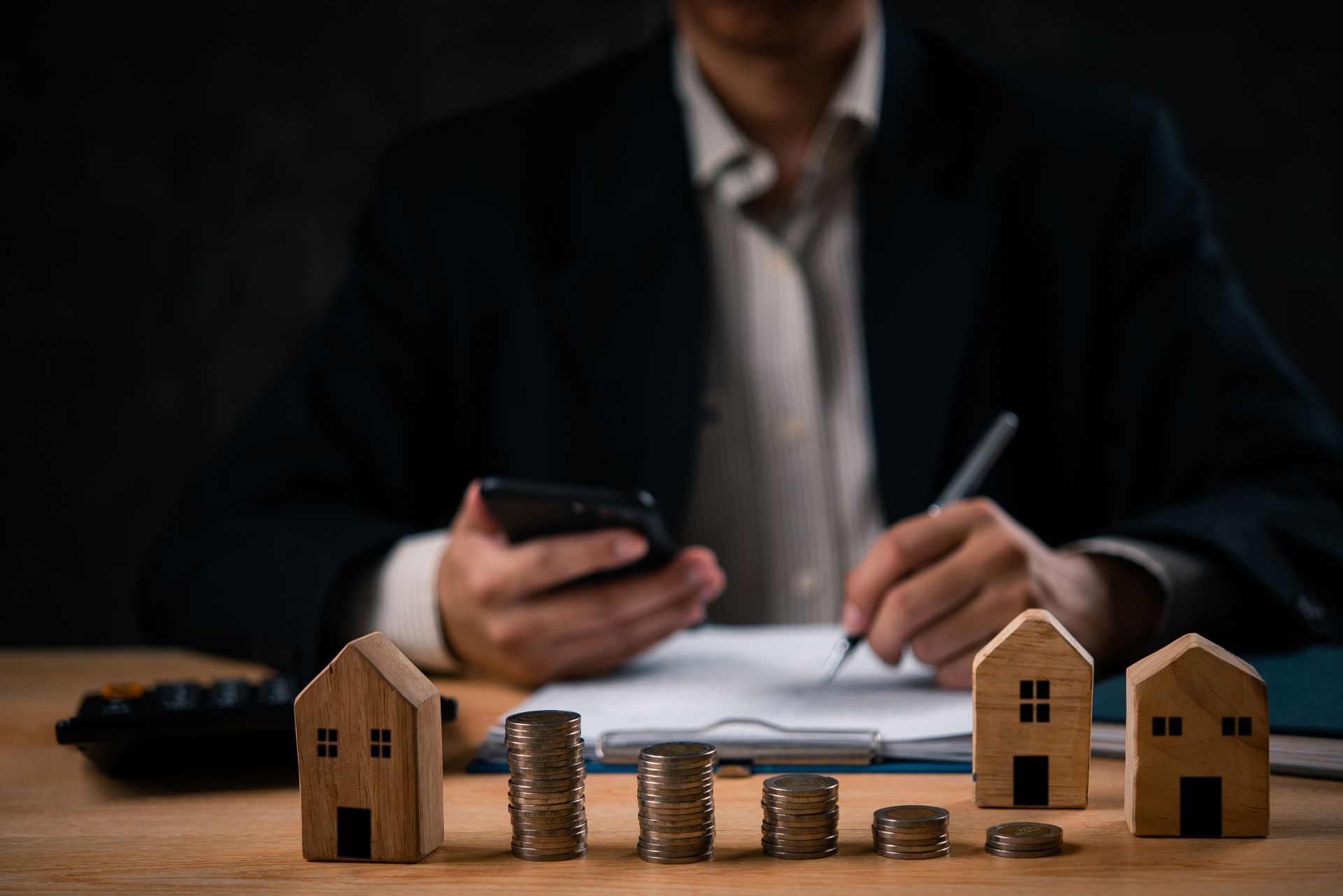 Person writing and using a phone behind small wooden houses and stacked coins on a table