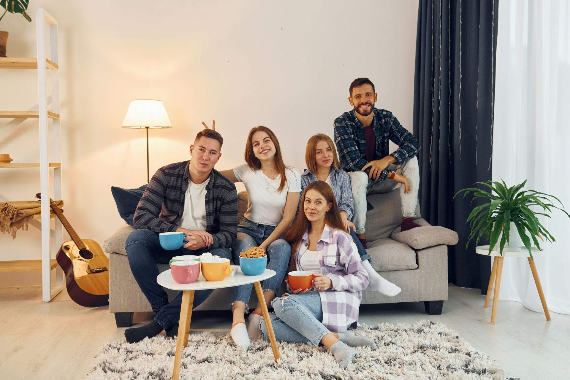 A group of four friends sits on a sofa and floor in a living room, smiling while holding mugs and bowls of snacks.