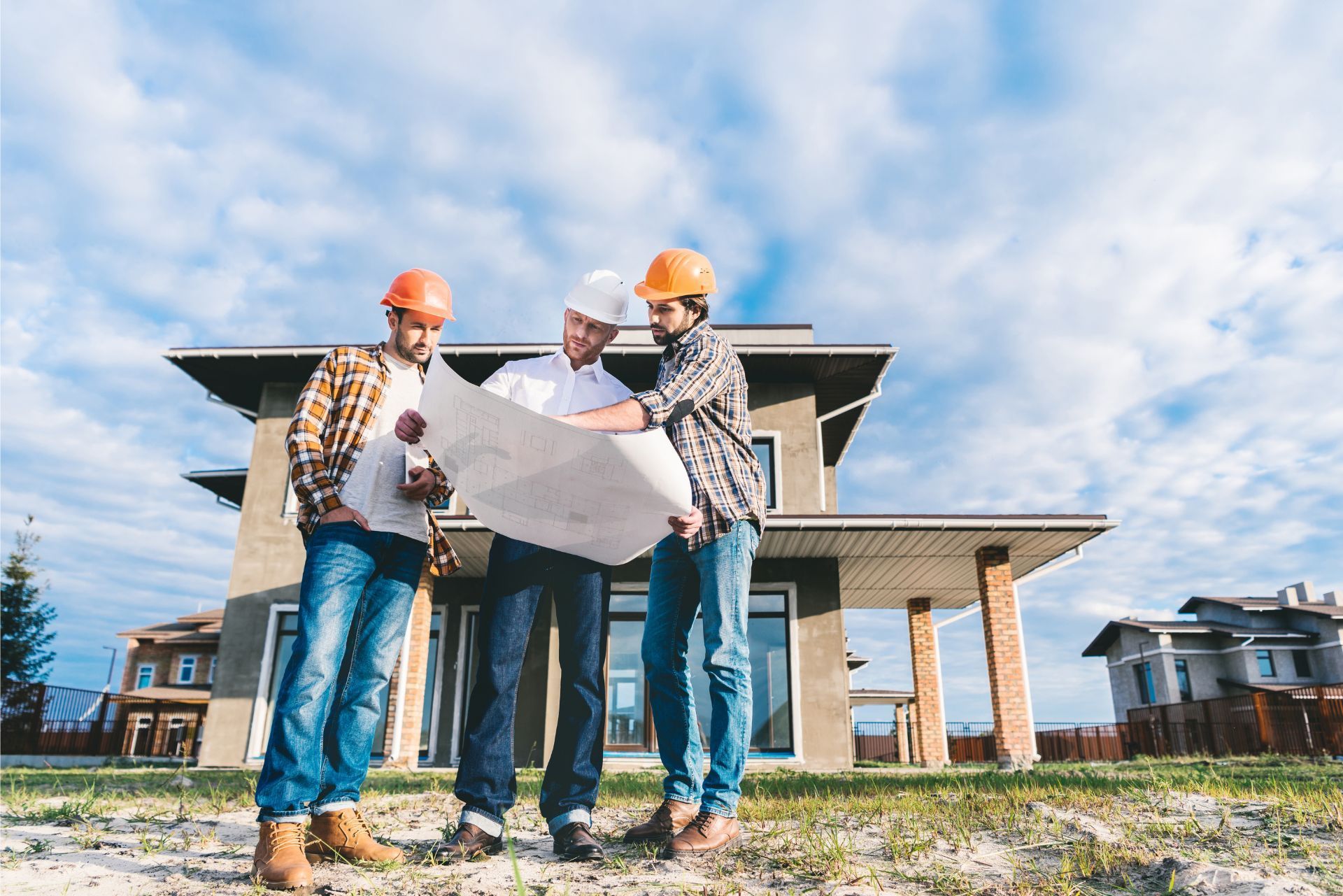 Three construction workers in hard hats reviewing blueprints outside a house under construction