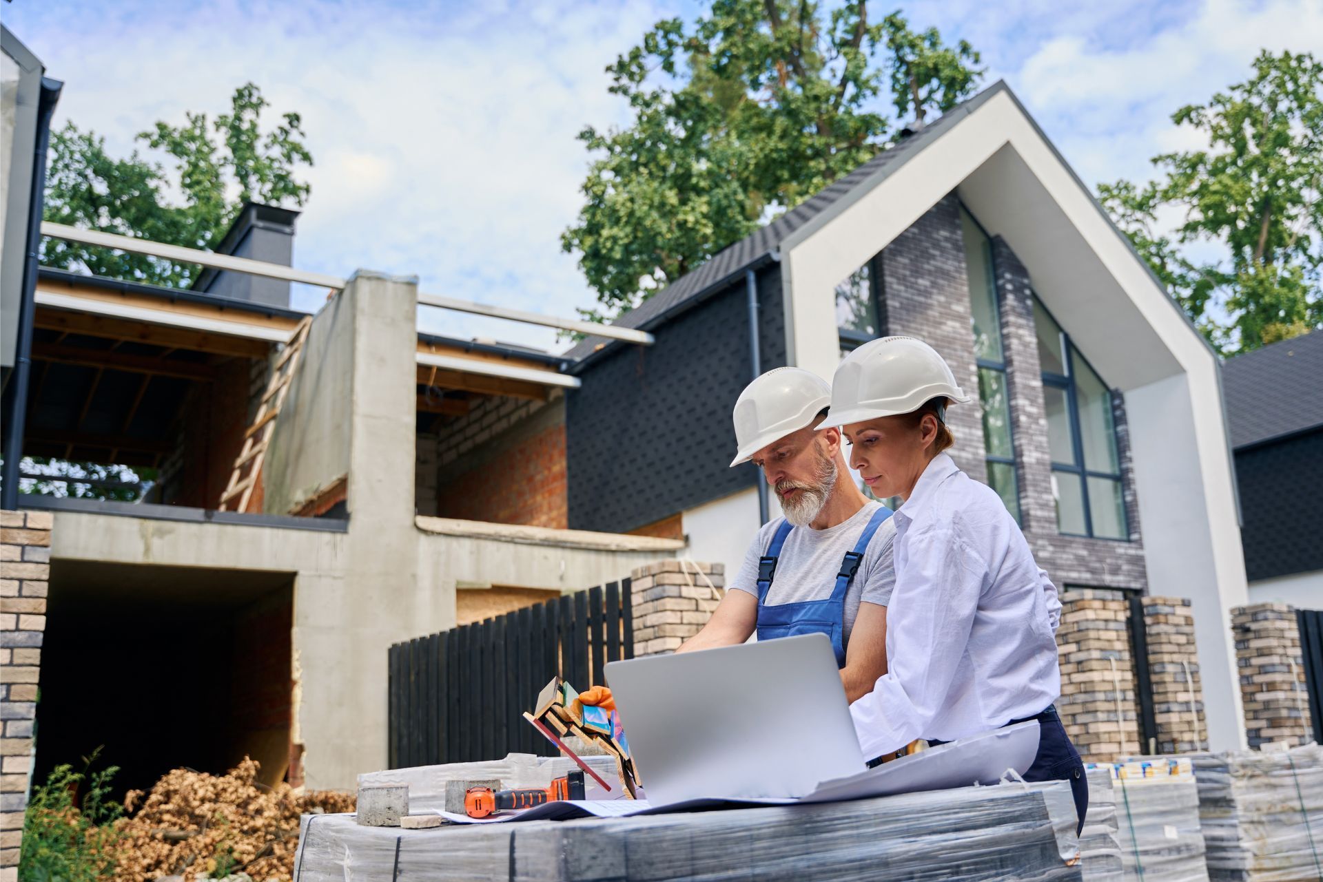 Two construction workers review plans on a laptop outside a house under construction.