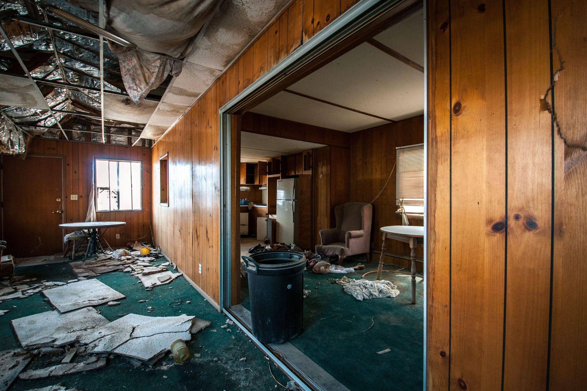 Abandoned, debris-filled room with wood-paneled walls, torn ceiling, and green carpet in disrepair