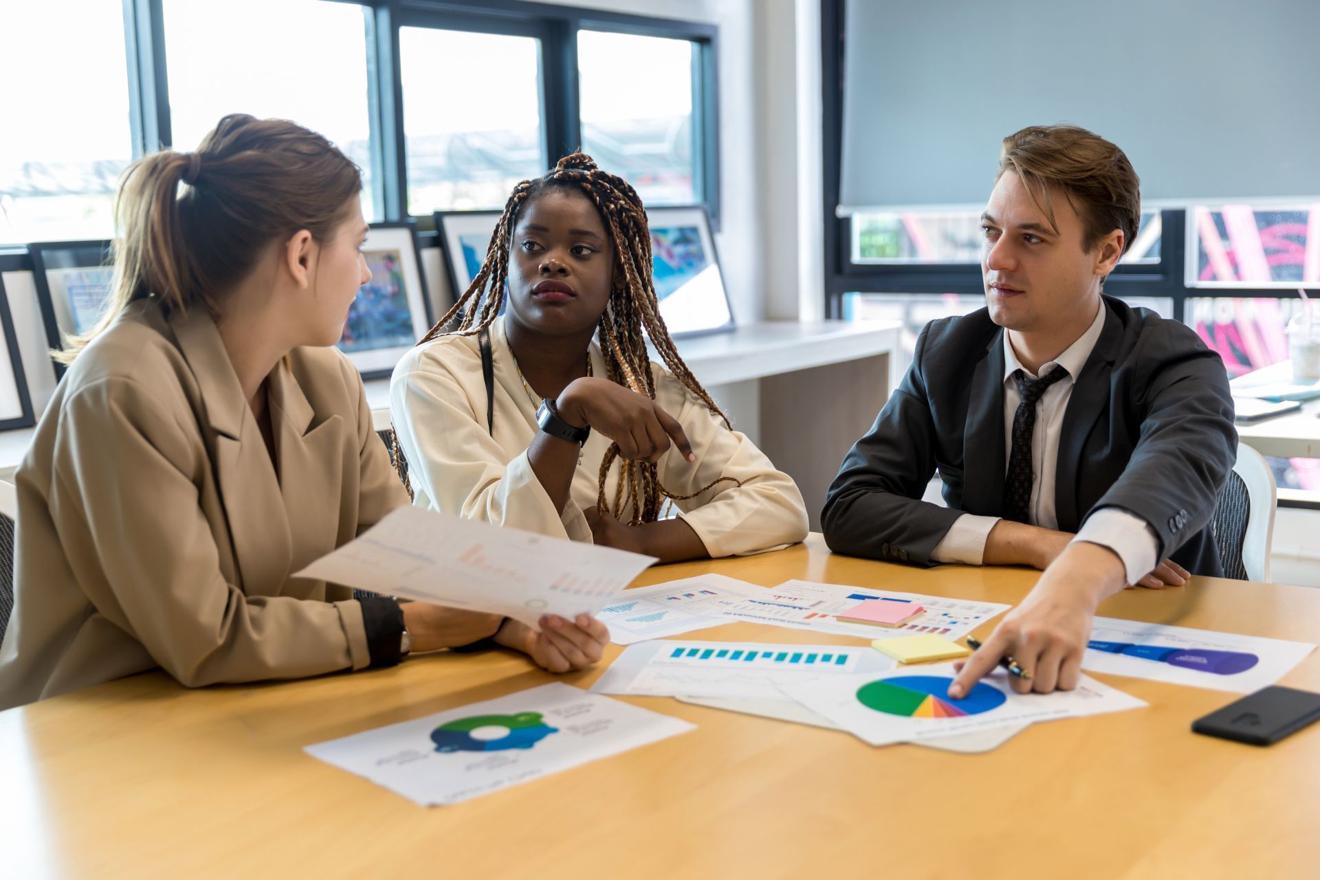 Three coworkers discussing printed charts at a table in a bright office room.