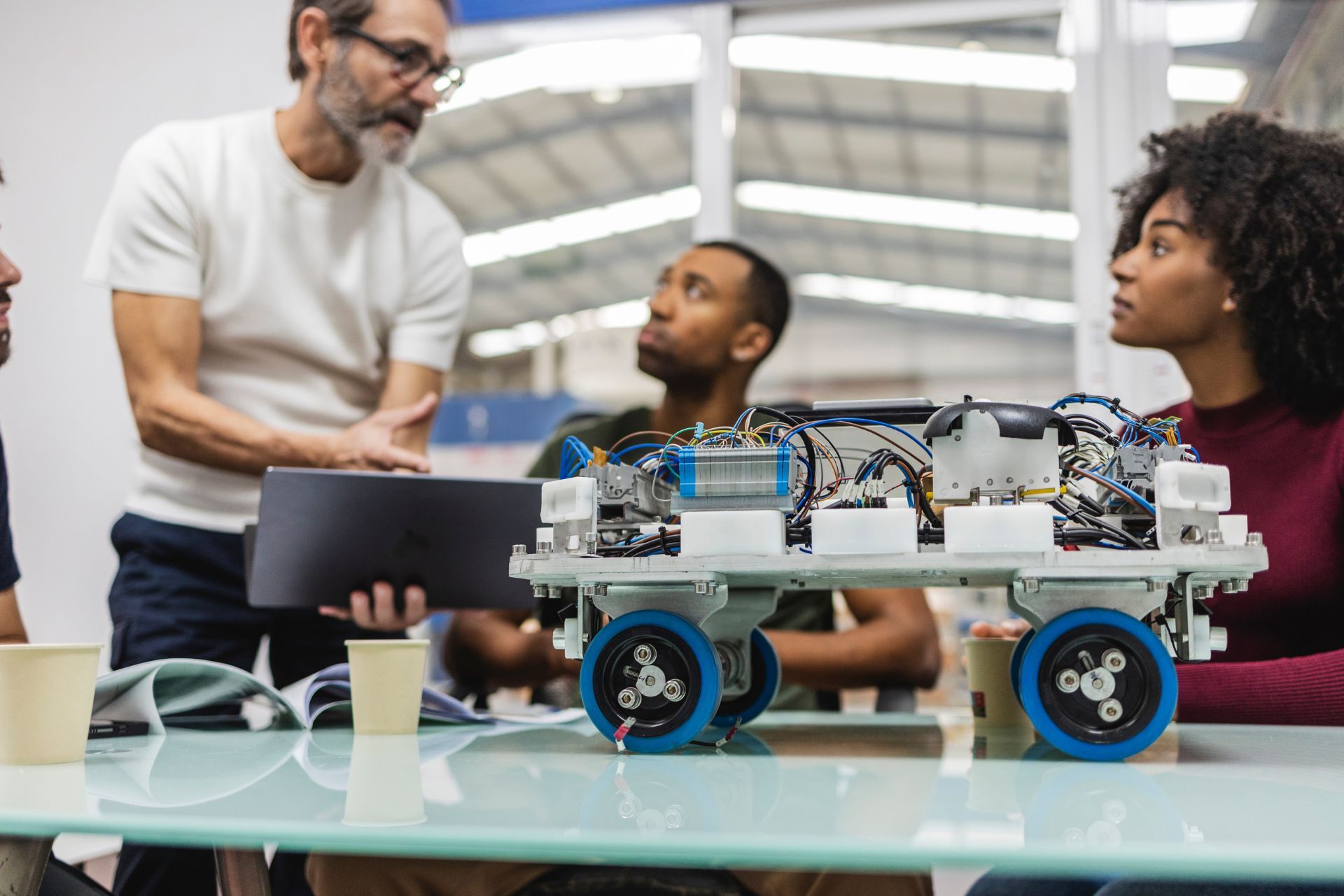 Three people gather around a table to discuss and examine a robotic chassis with exposed electronics and blue wheels.