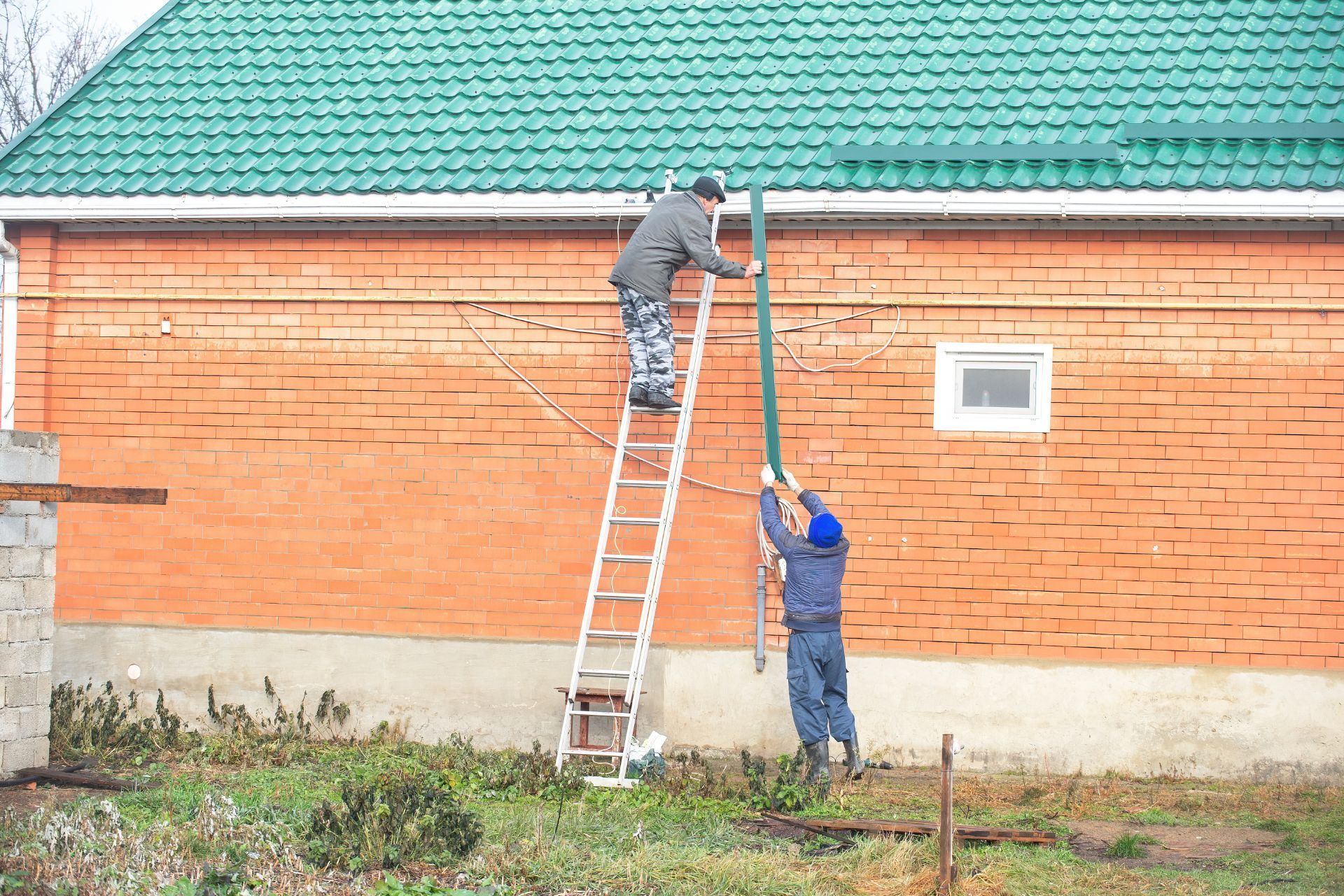 Two workers on ladders repair orange brick wall beneath a green roof