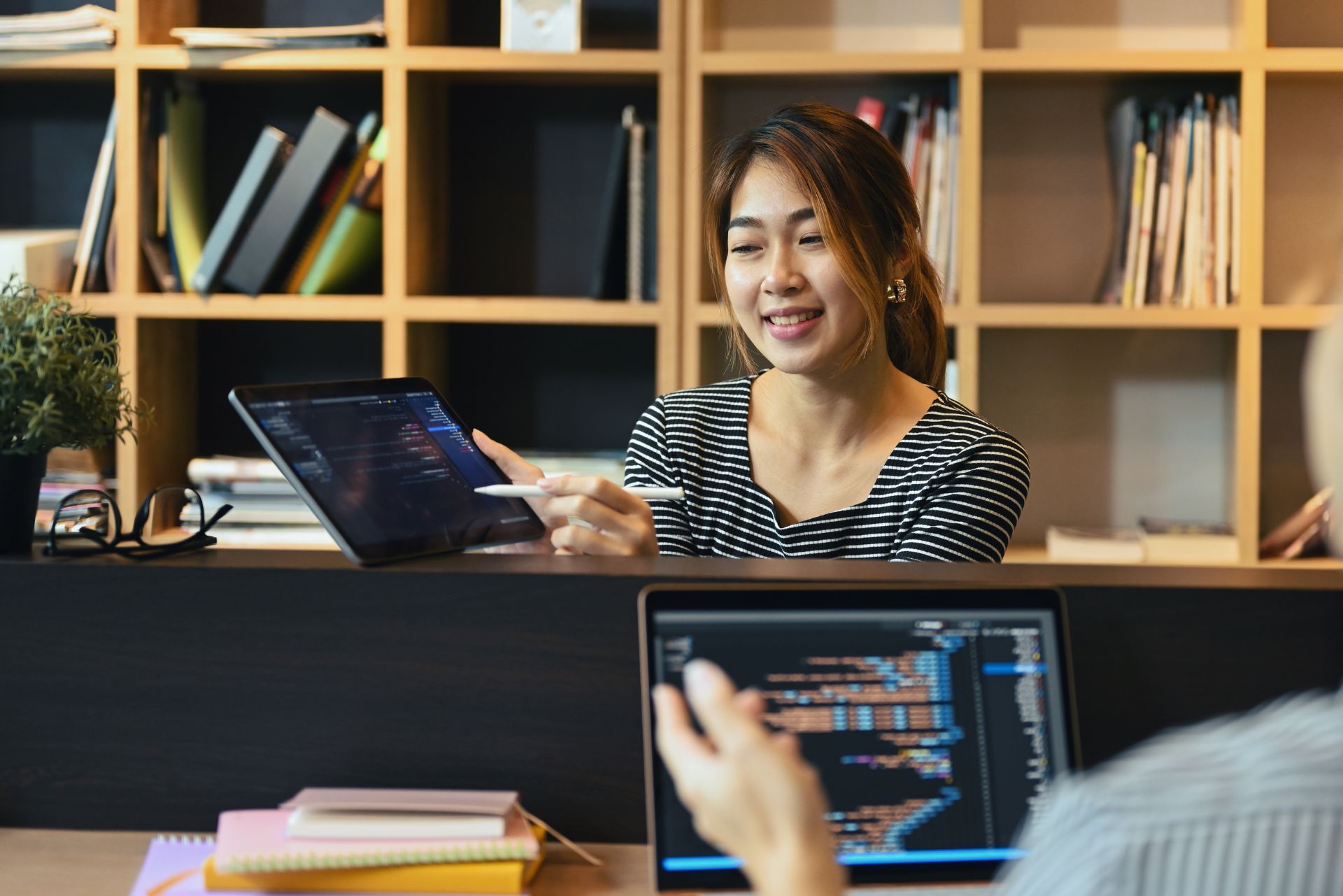 Woman smiling at a laptop in an office, with shelves behind her and another laptop in the foreground