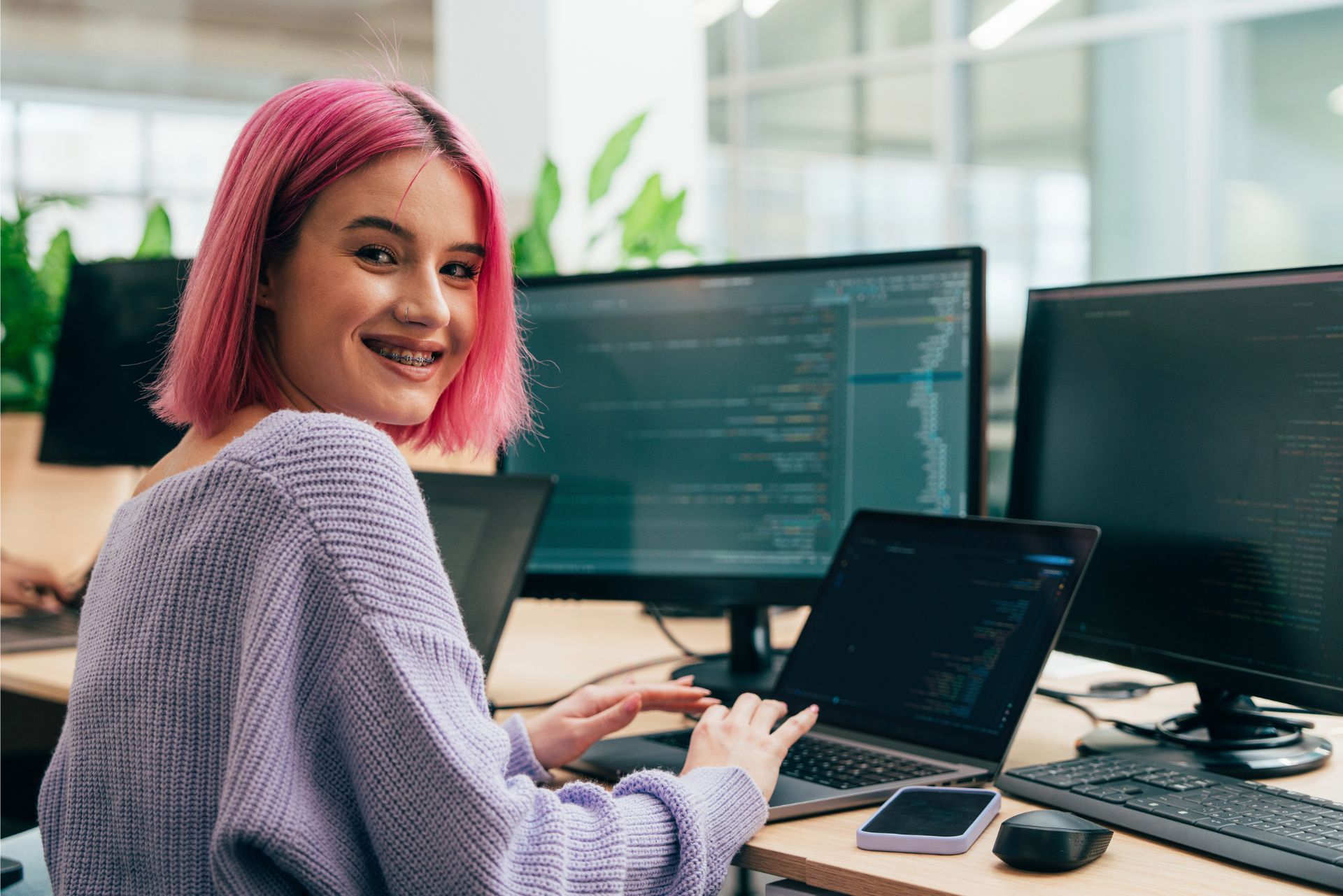 Person with pink hair smiling at a desk with multiple monitors in an office