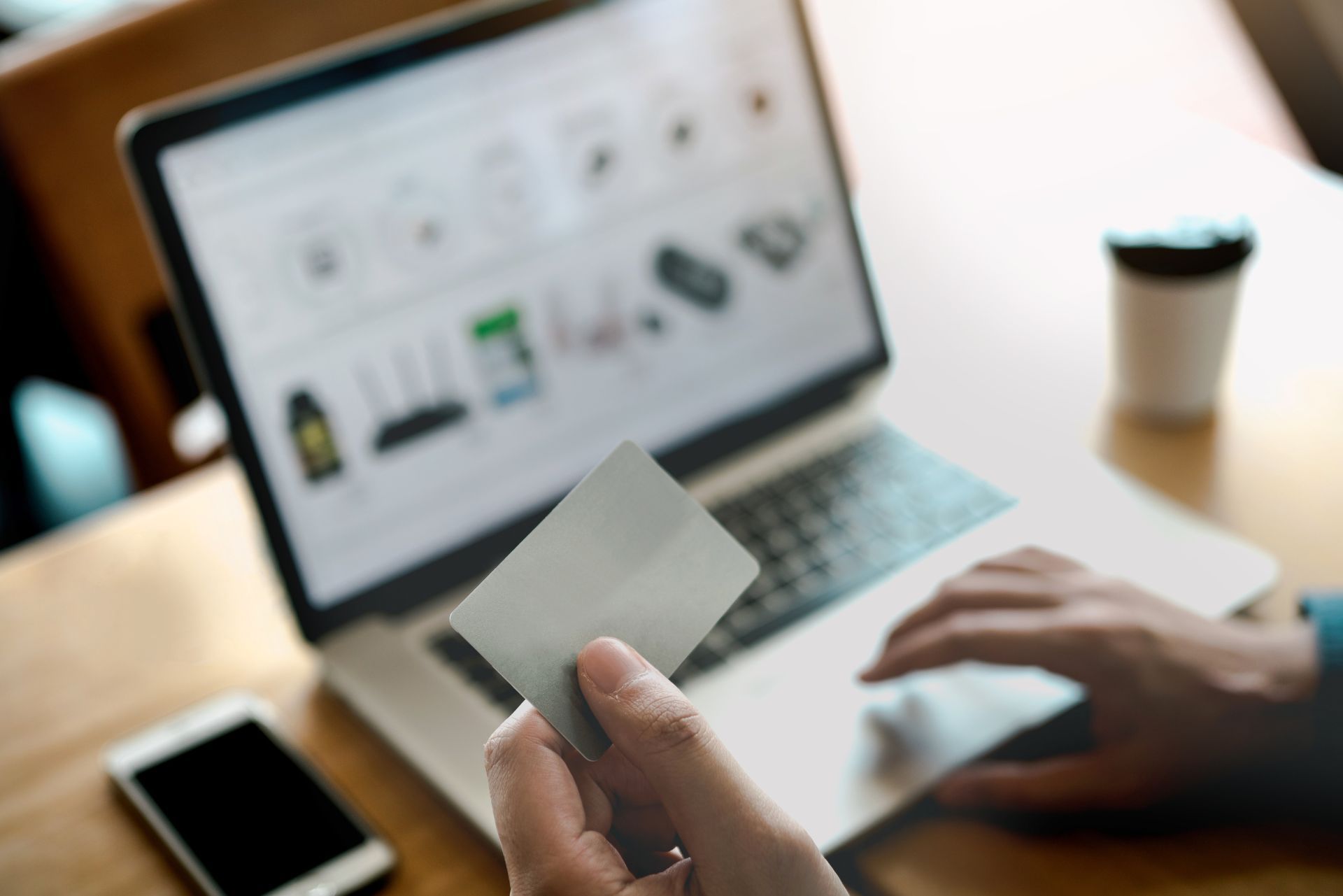 A person holding a credit card while shopping on a laptop at a wooden table with a smartphone and a coffee cup.