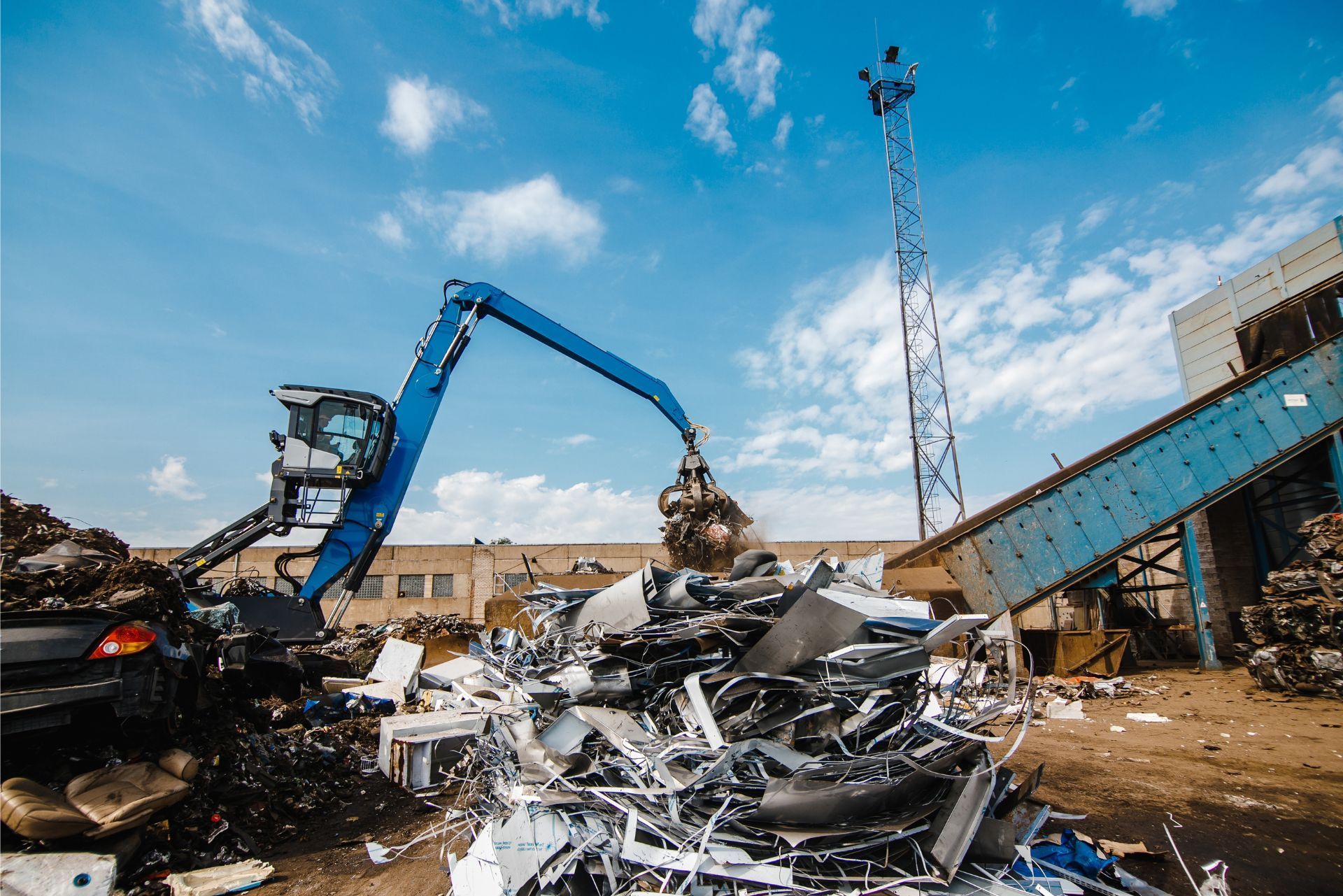 Blue excavator sorting scrap metal in a junkyard under a clear sky