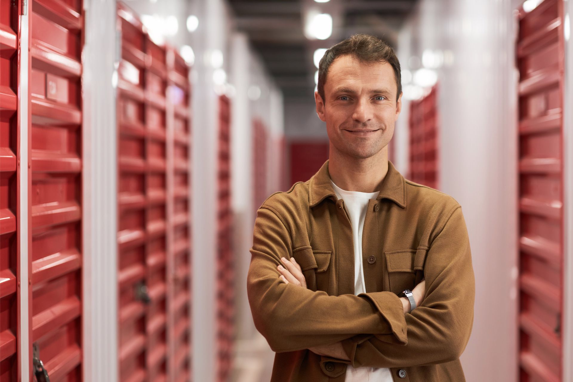 Man standing with arms crossed in a storage aisle between red shelves, wearing a brown jacket.