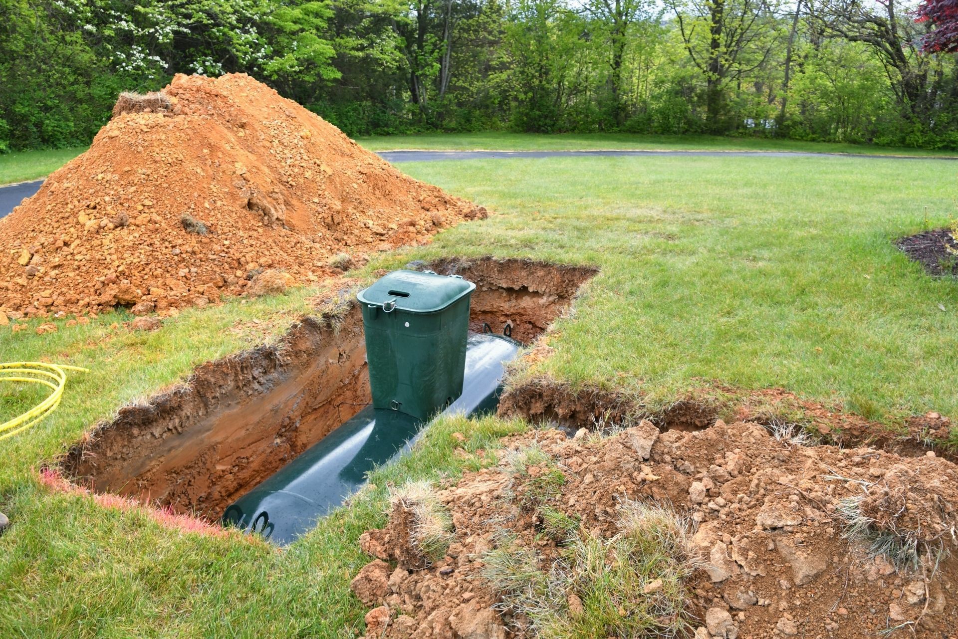 A large green septic tank partially installed in a trench in a grassy yard next to a pile of excavated dirt.