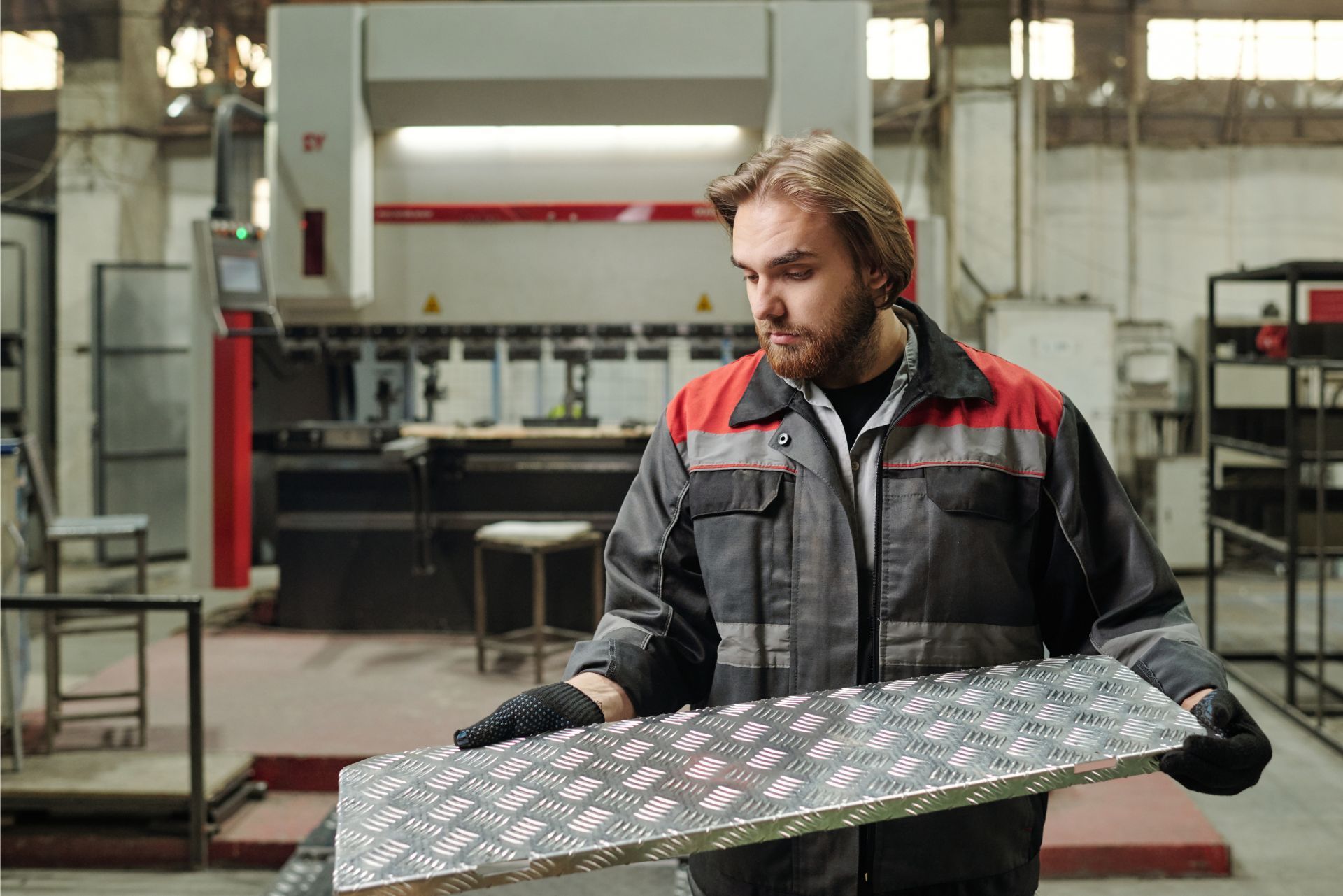 Worker in a factory holding a large metal sheet, wearing a gray and red work jacket.