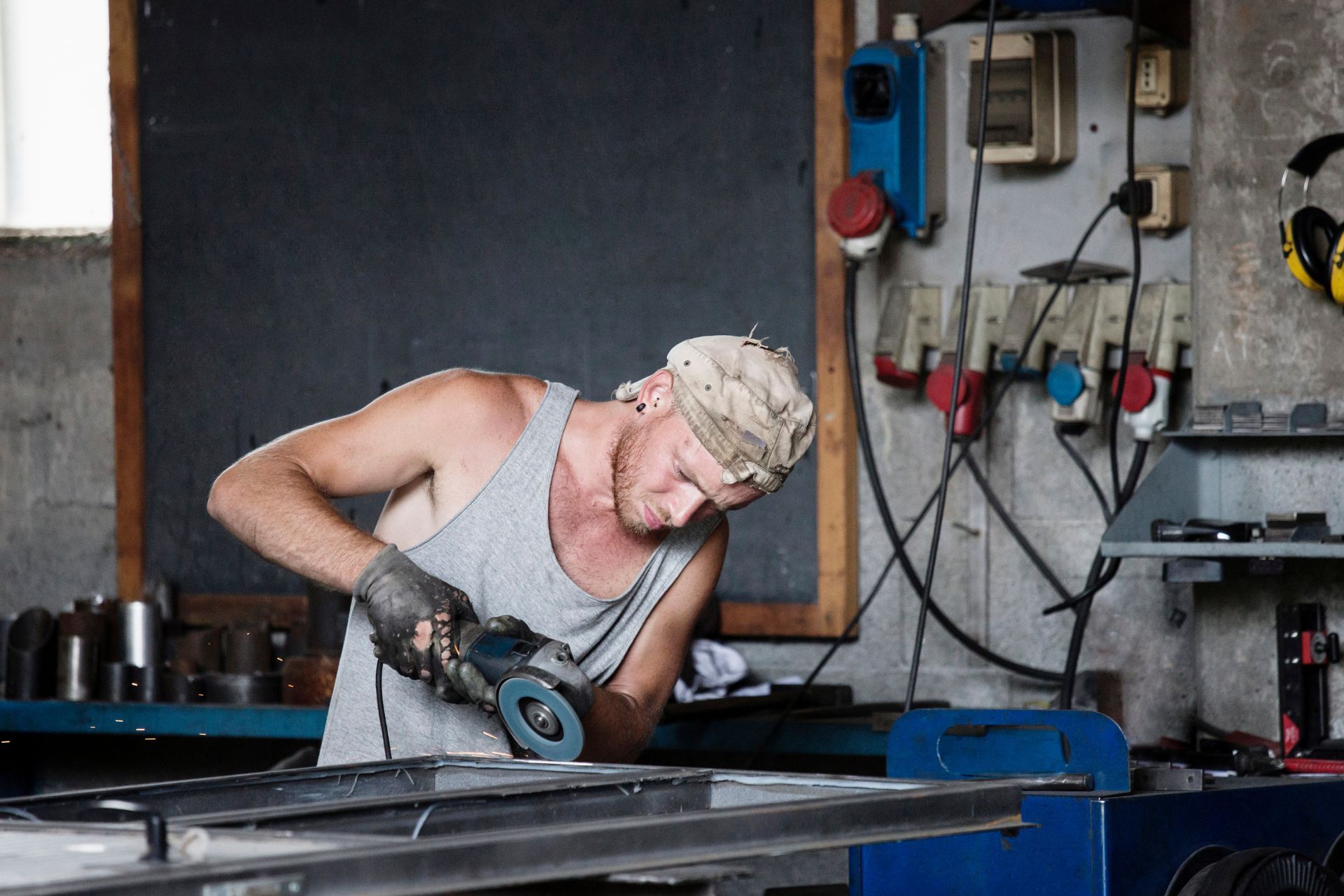 Worker using a power tool on a metal sheet in a workshop, with equipment mounted on the wall.