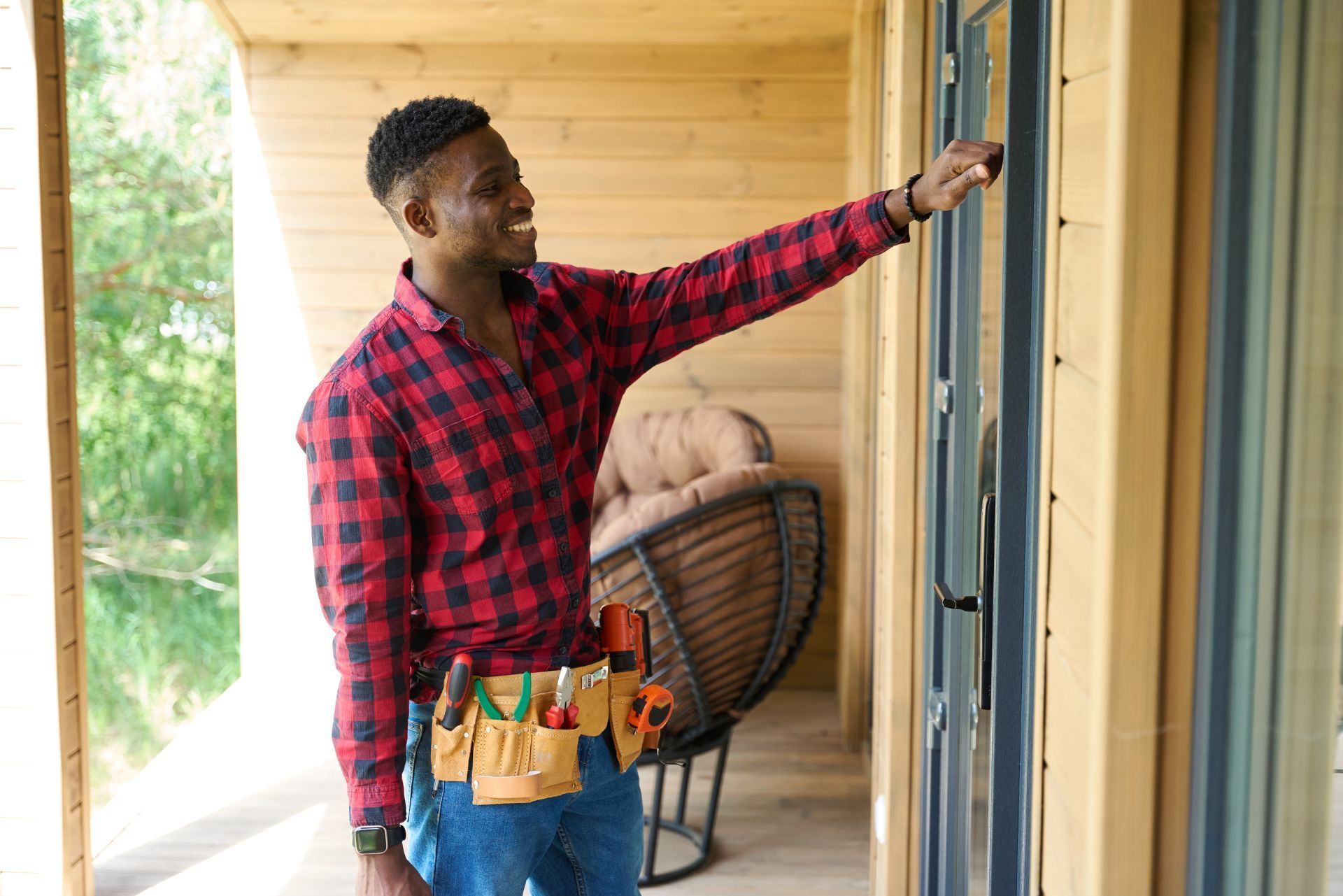 Man in a red plaid shirt hanging a door frame on a wooden porch with tools at his waist