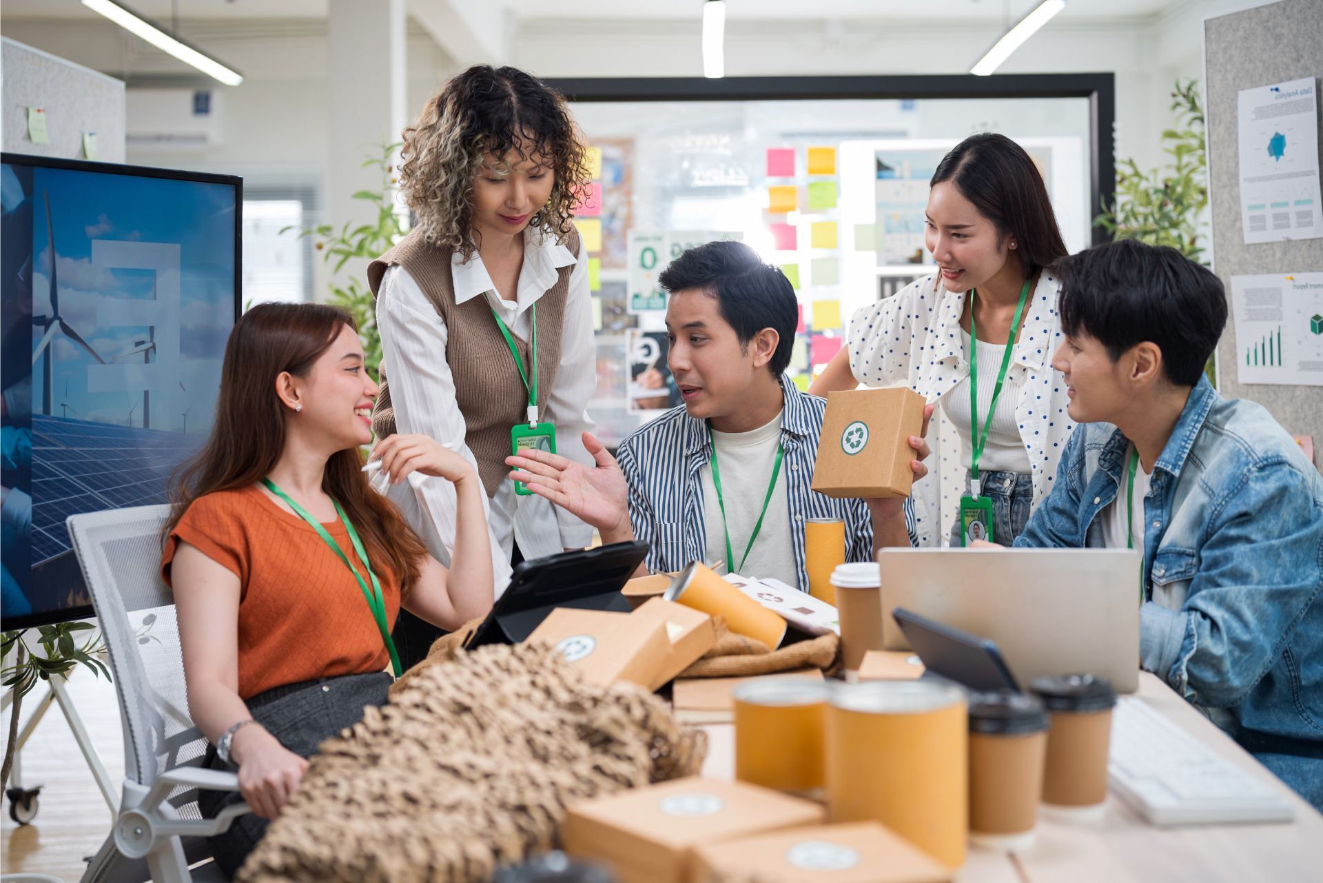 Team collaborating around a laptop in a bright office with notes, coffee cups, and sticky notes on the wall