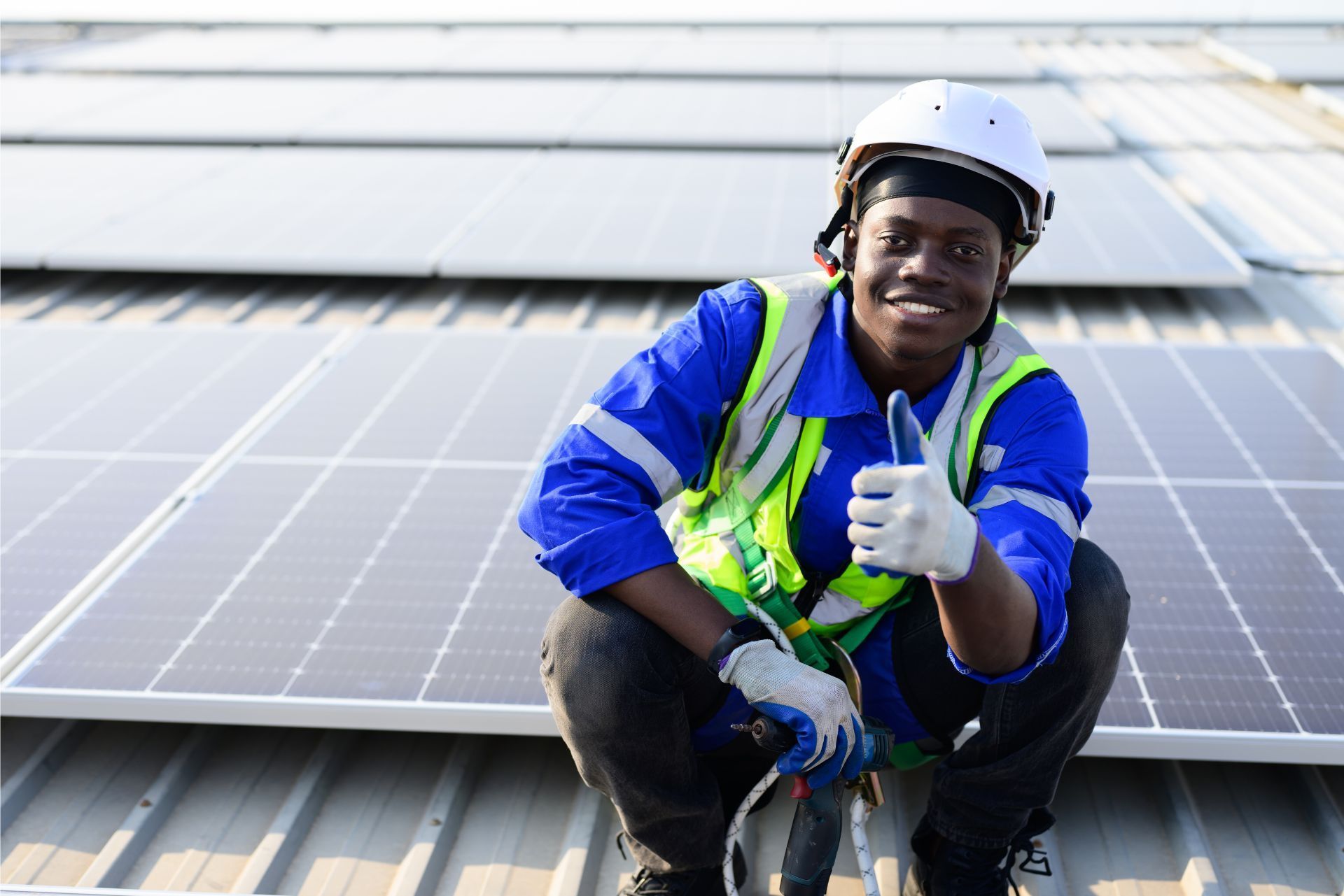 Worker in safety gear kneels by solar panels and gives a thumbs-up on a rooftop.