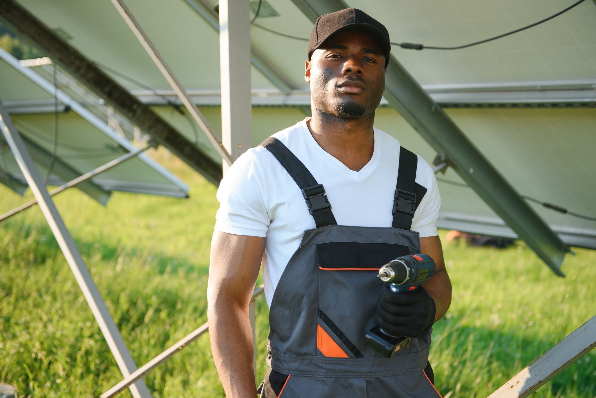Man standing in black overalls and cap beside a large solar panel in a grassy field, holding a camera
