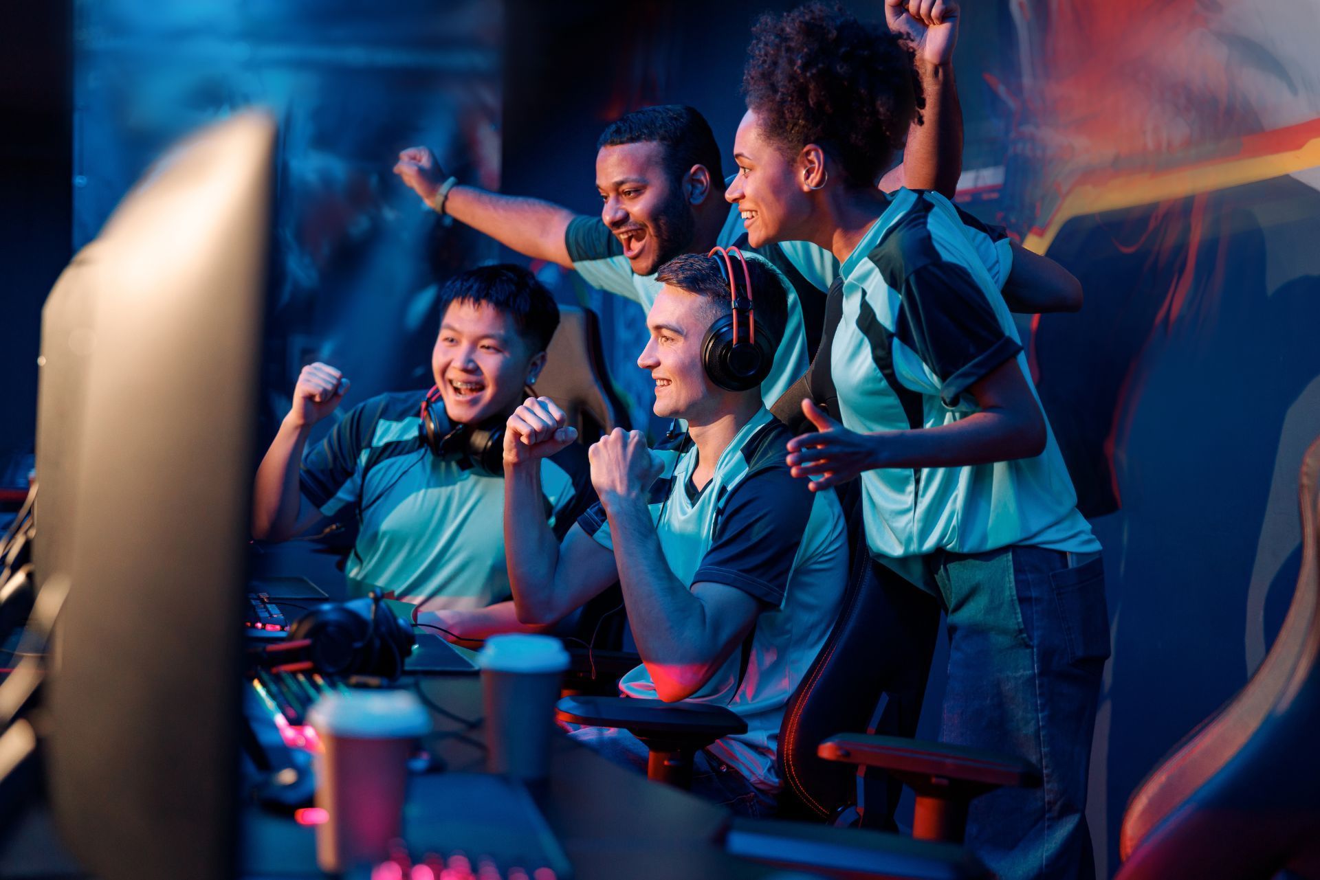 Four teammates in teal jerseys cheering at gaming stations with neon lights