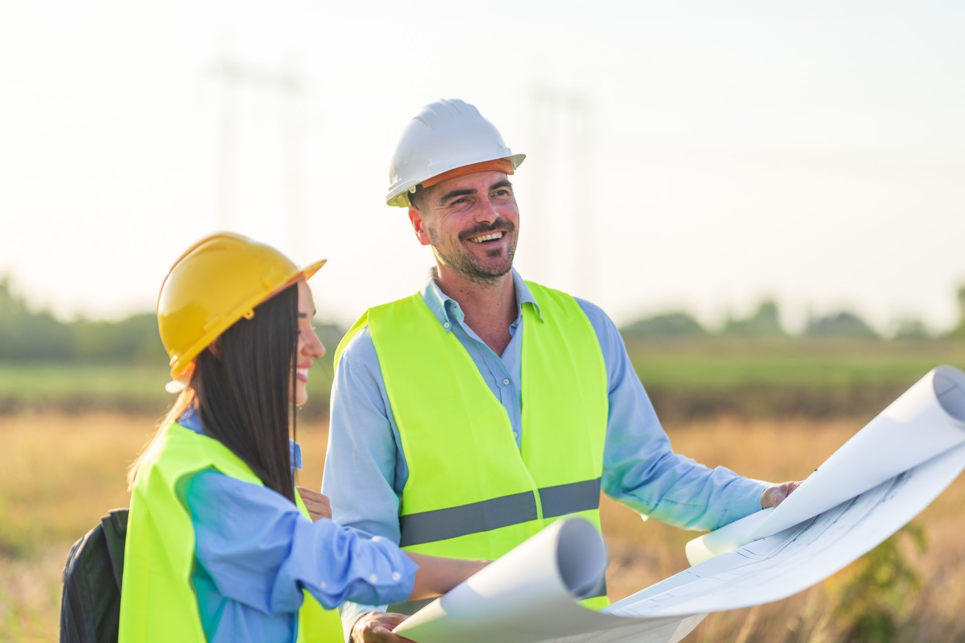 Two people in safety vests and hard hats look at construction blueprints outdoors in a field.