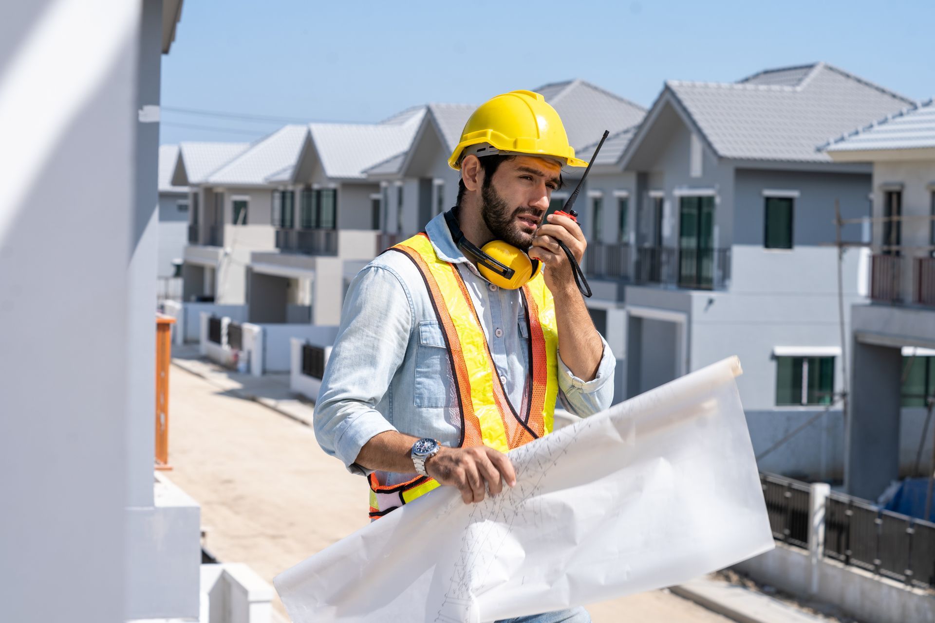 A construction worker in a hard hat and safety vest holds blueprints while speaking into a radio at a housing site.