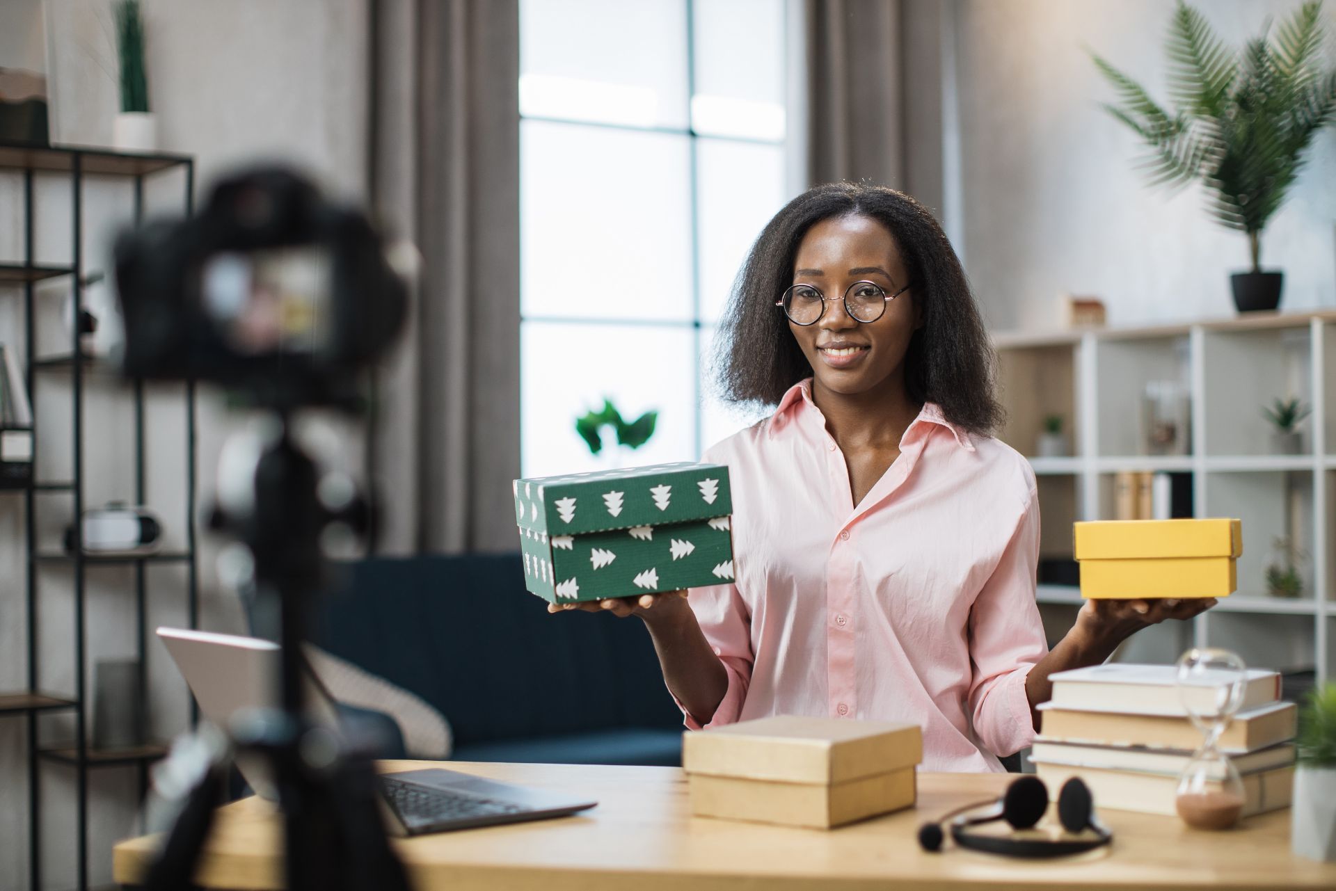 A person sits at a desk in a bright room, holding a green box and a yellow box while recording a video on a camera.