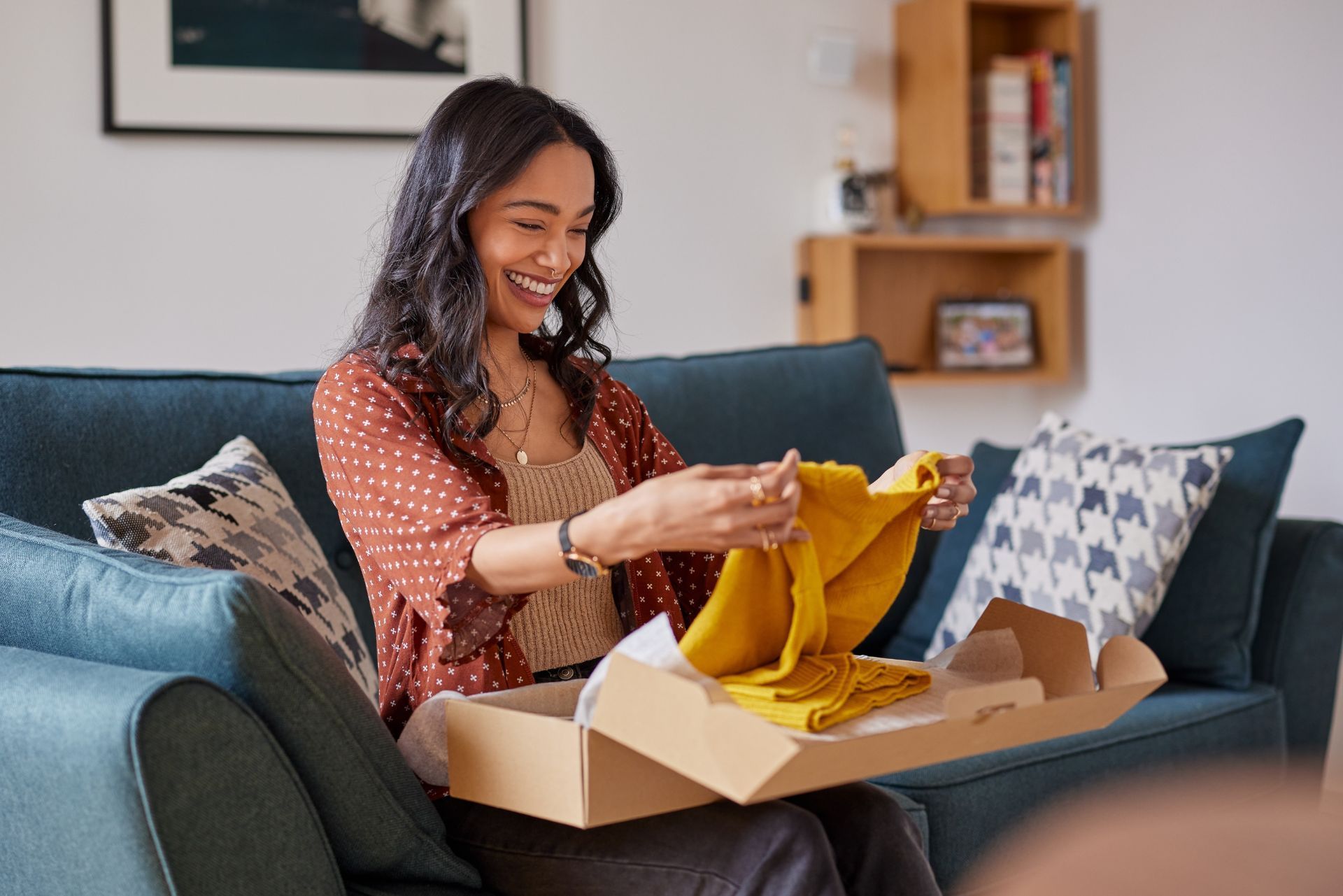 A person smiling while sitting on a couch, holding a yellow garment taken from a cardboard shipping box.