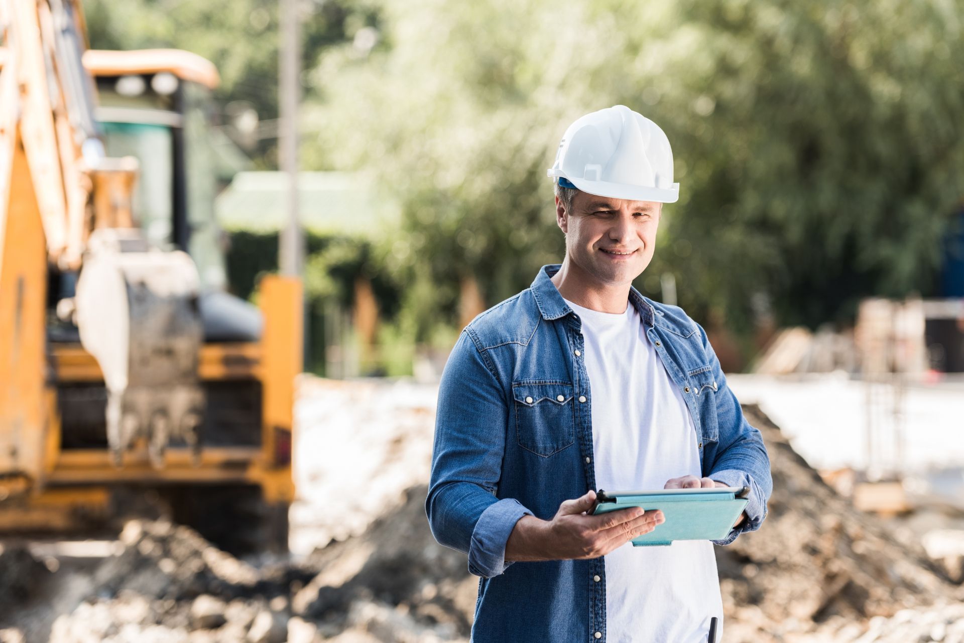 A smiling construction worker wearing a white hard hat holds a tablet on a sunny, dirt-filled job site with a yellow excavator.