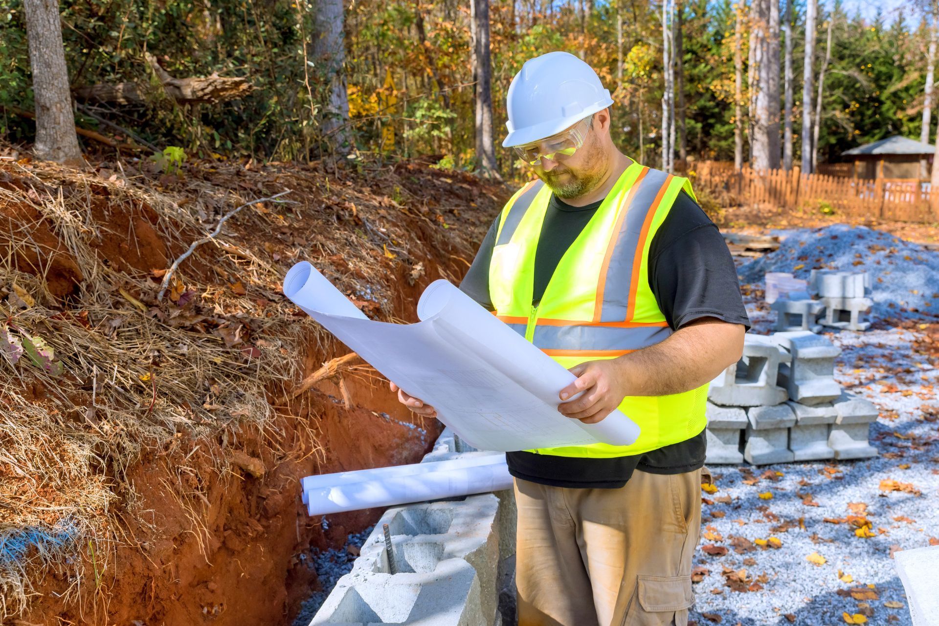 Construction worker in a safety vest and hard hat reviewing blueprints at an outdoor job site with concrete blocks.