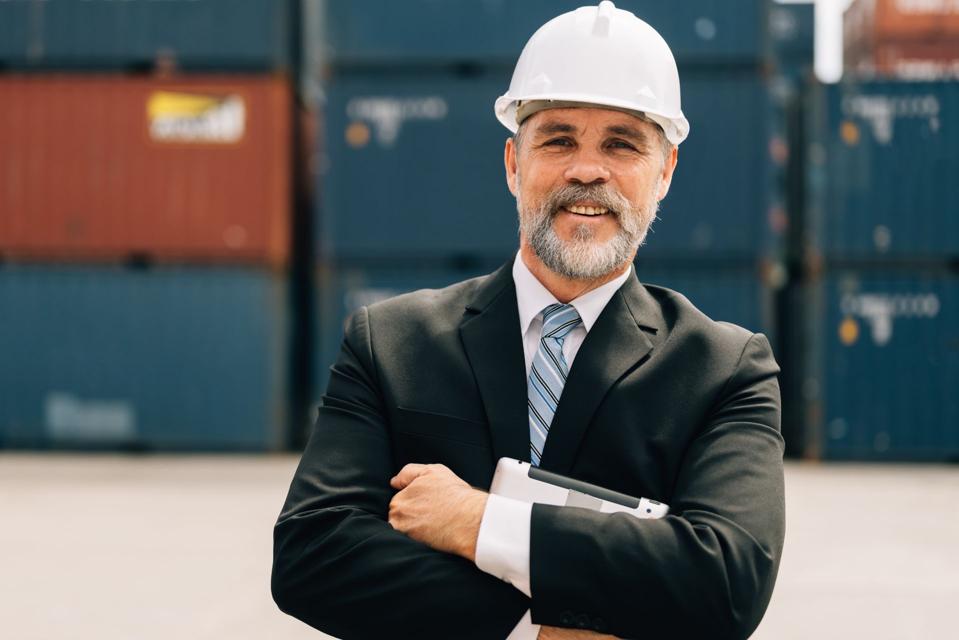 A professional wearing a white hard hat and suit smiles with arms crossed, standing before stacked shipping containers.
