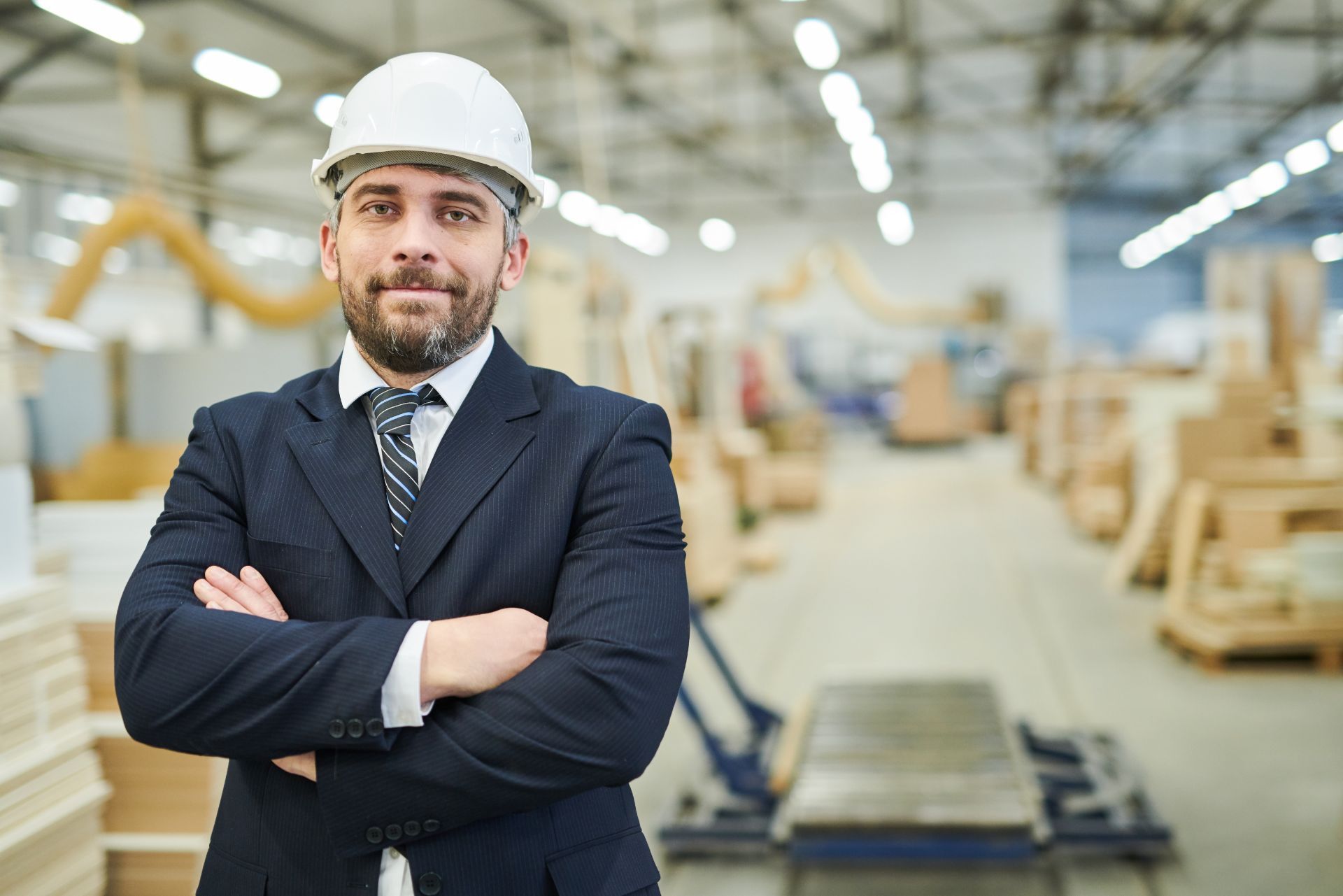 A person wearing a white hard hat and suit with arms crossed, standing in a blurred industrial warehouse facility.