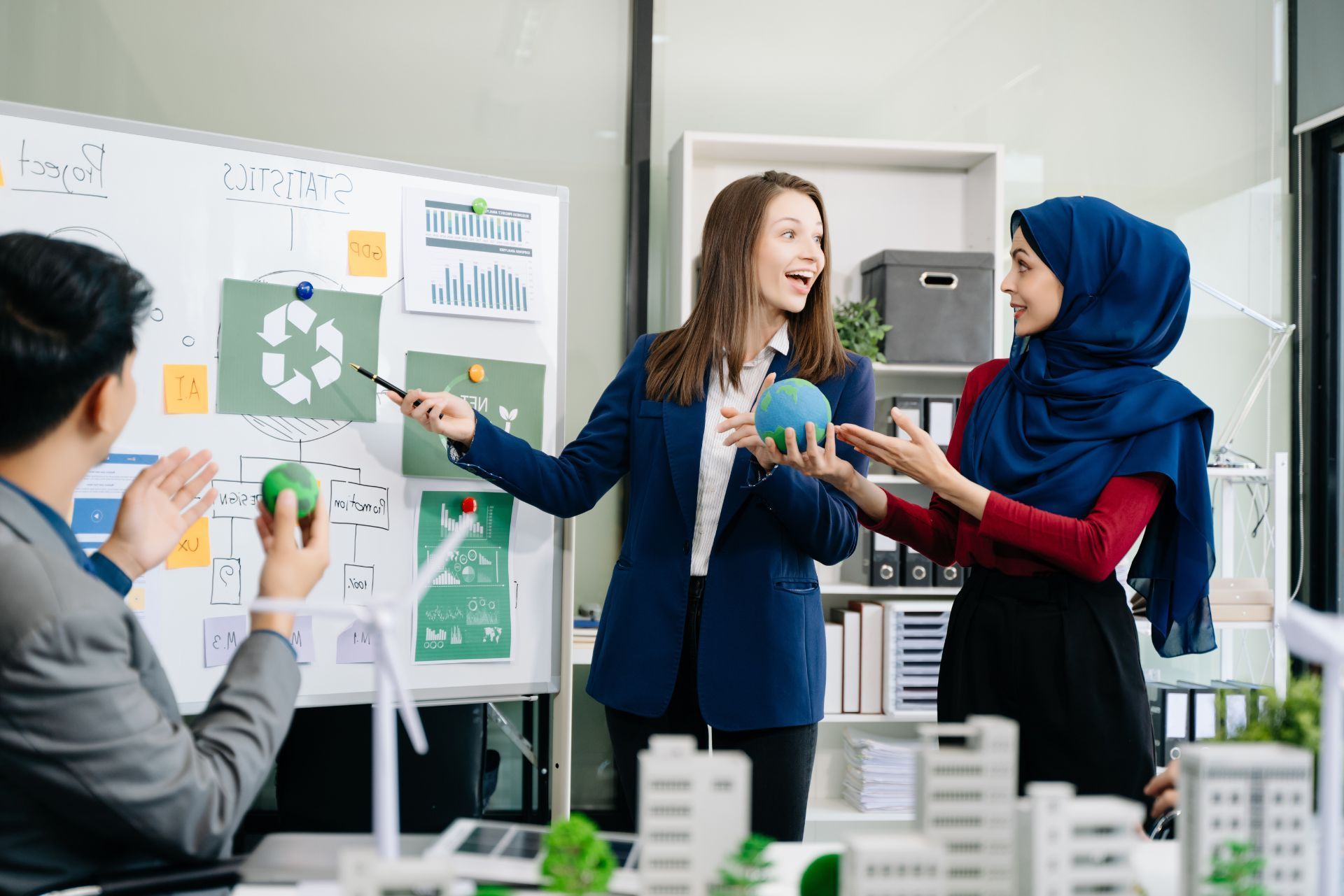 Three professionals collaborate in an office, discussing sustainability projects using a whiteboard and building models.
